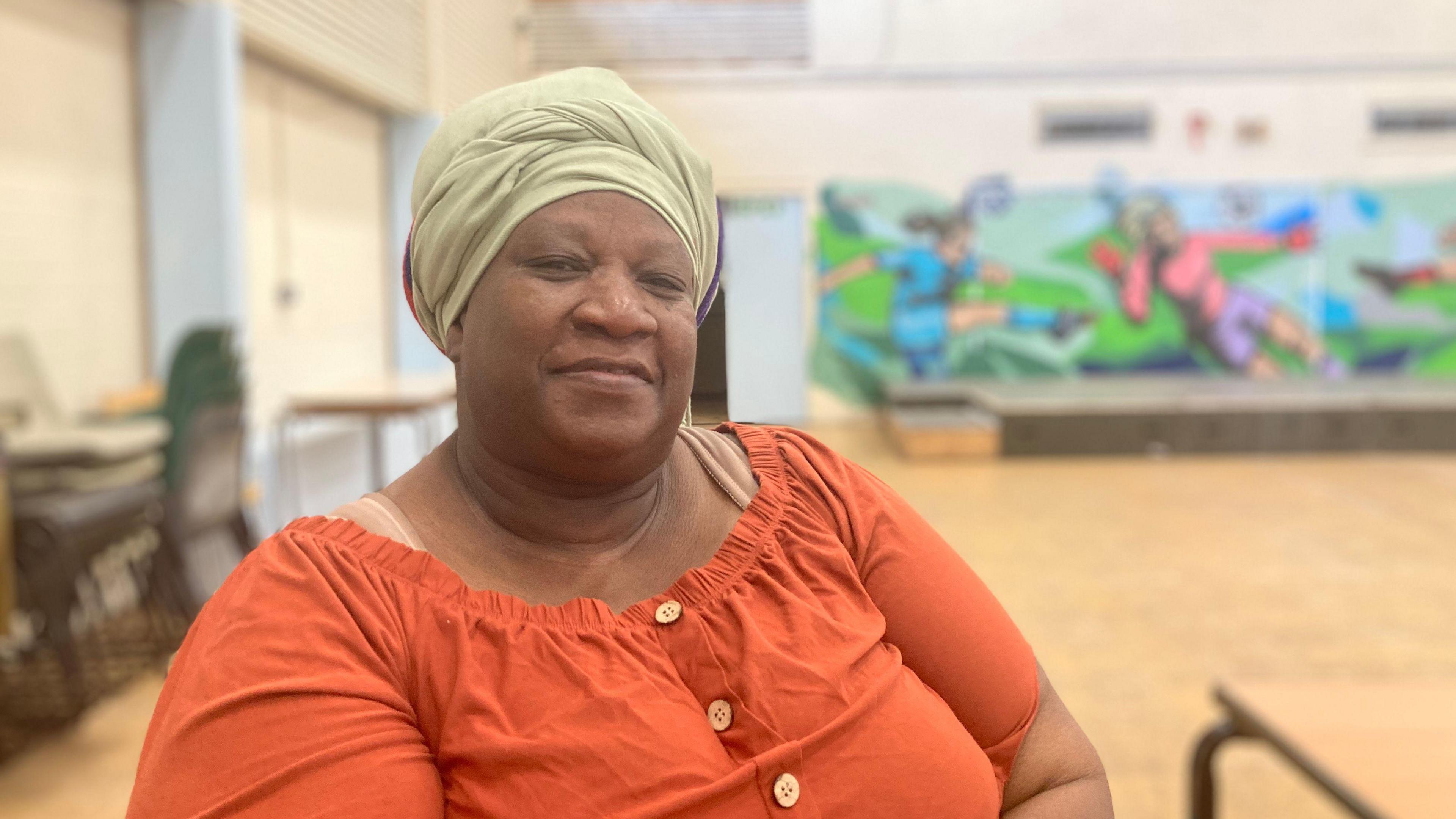 A woman, smiling - she has a sage green scarf around her head and a bright orange top with buttons. She is sitting in a community centre.