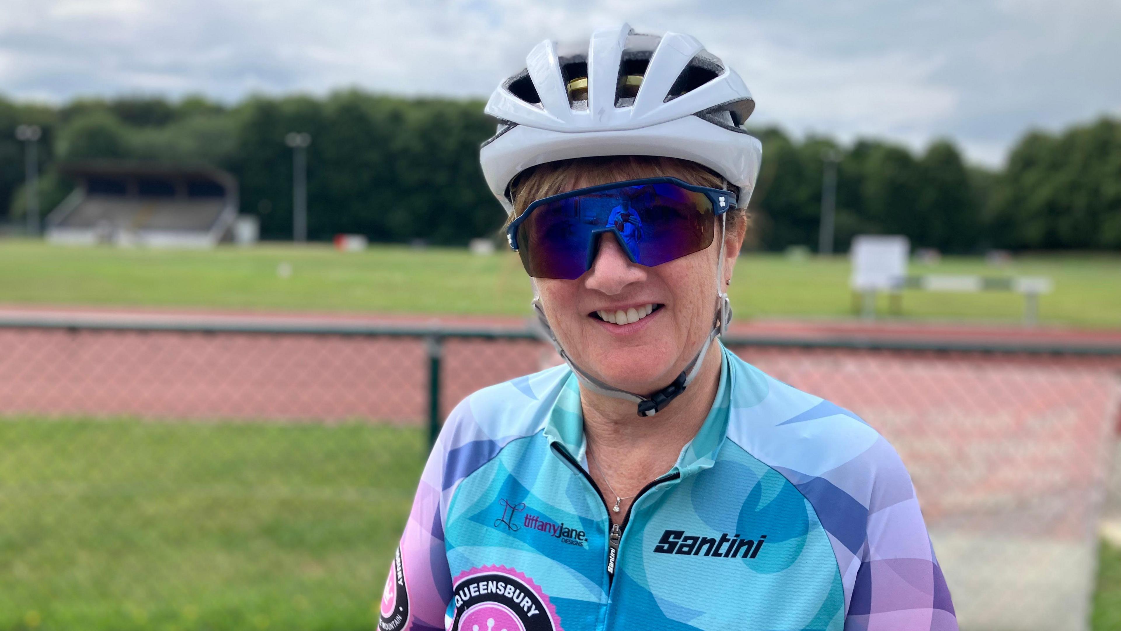 A woman wears a white cycling helmet and cycling sunglasses. She smiles at the camera as a race track is blurred behind her.