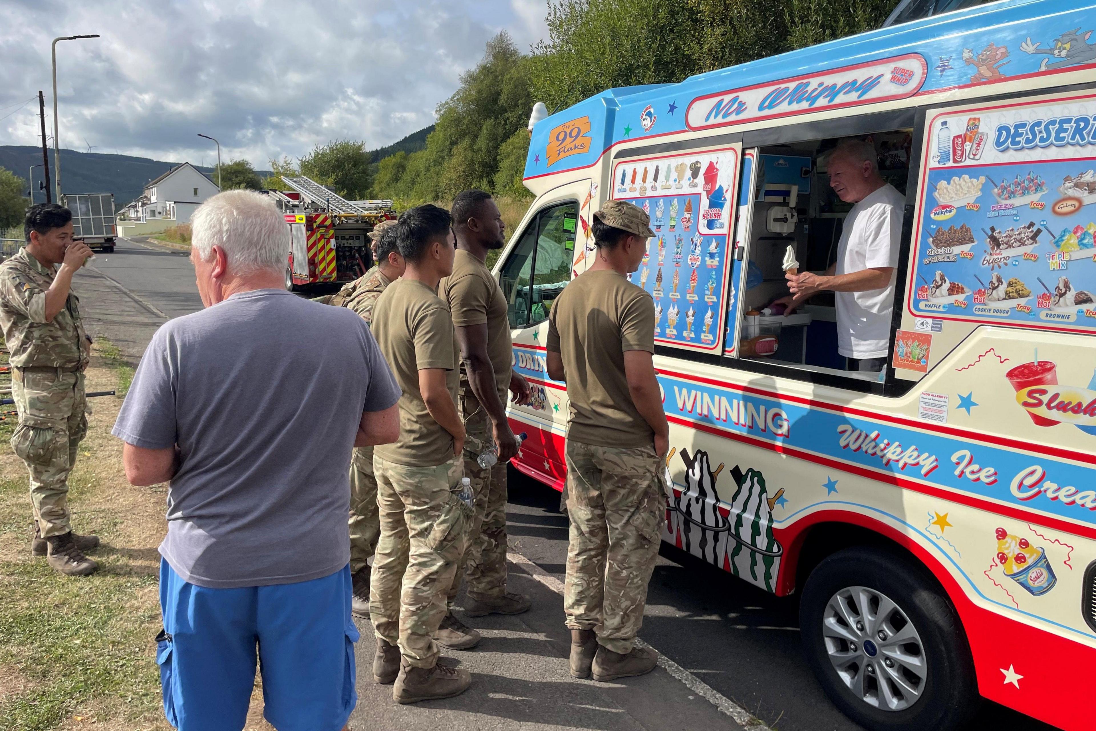 A group of soldiers are stood in a queue towards an ice cream van. In the van is a man holding out an ice cream.