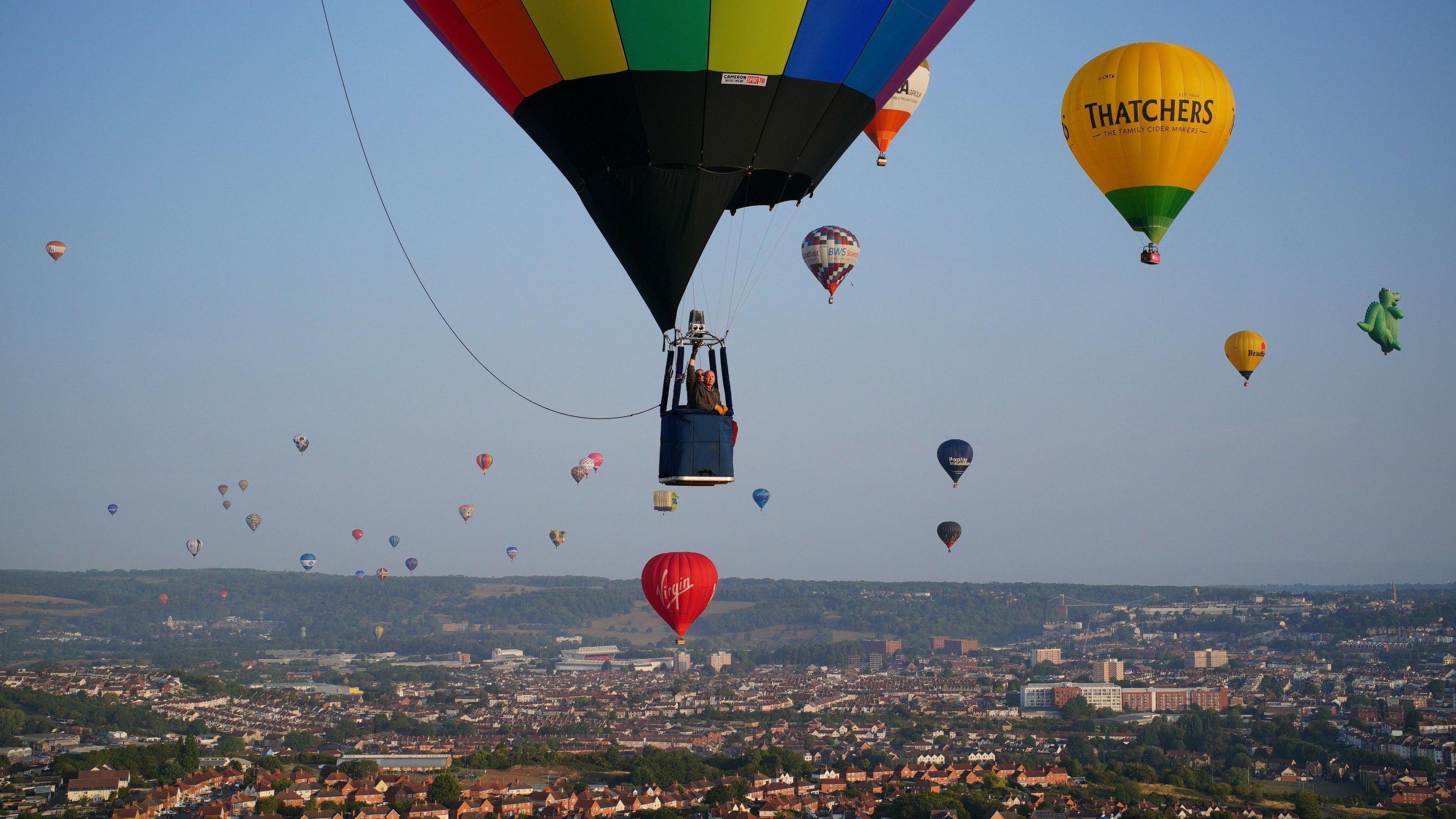 A pilot stands inside a basket as he flies a hot air balloon with a striped, rainbow-themed canopy. The basket is small and it appears he is the only person in it. Dozens of other balloons can be seen in the distance. They are all flying above Bristol, with the rooftops of houses visible.