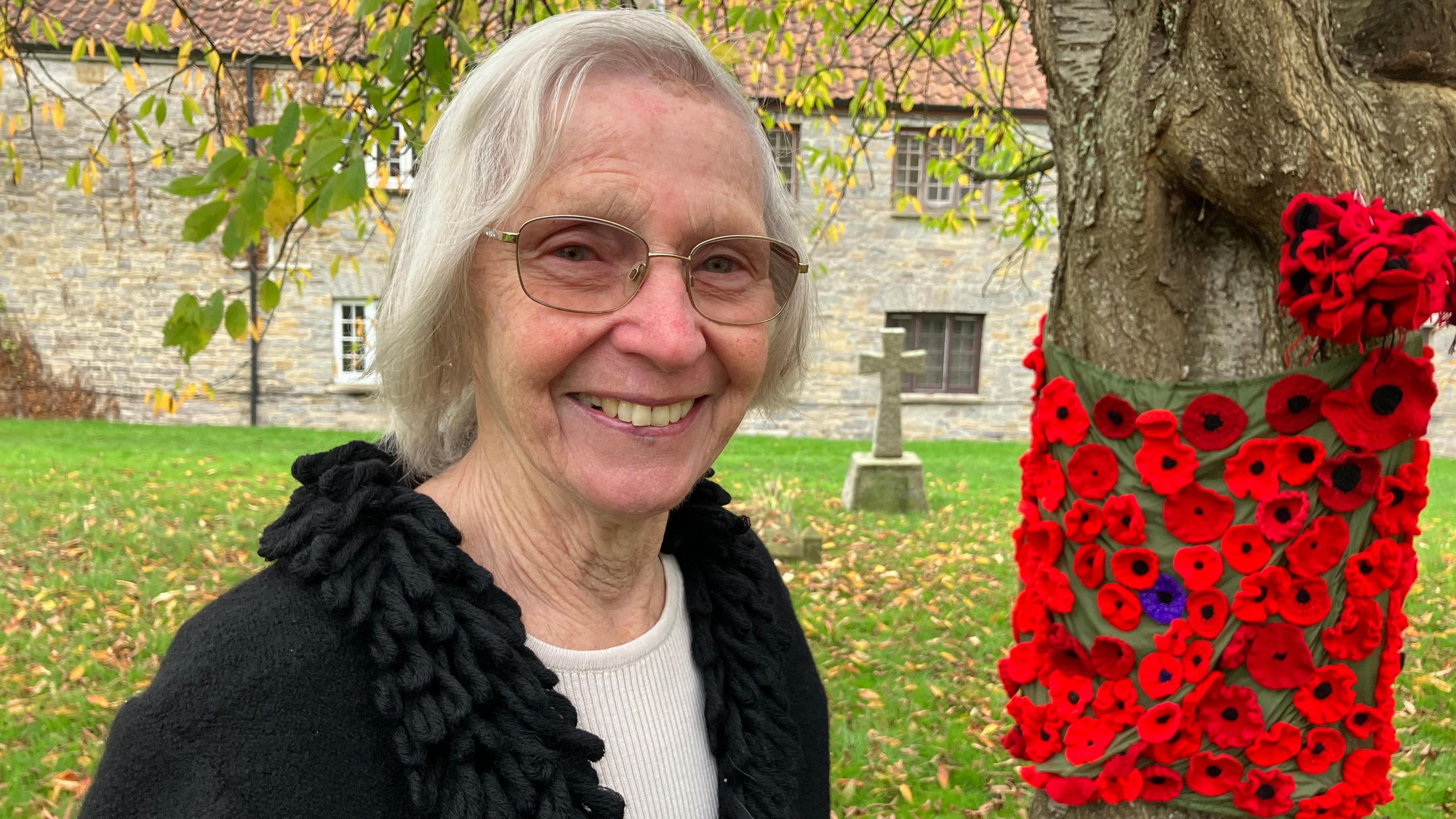 A women with grey hair and glasses looking at the camera with poppies attached to the trunk of a tree next to her