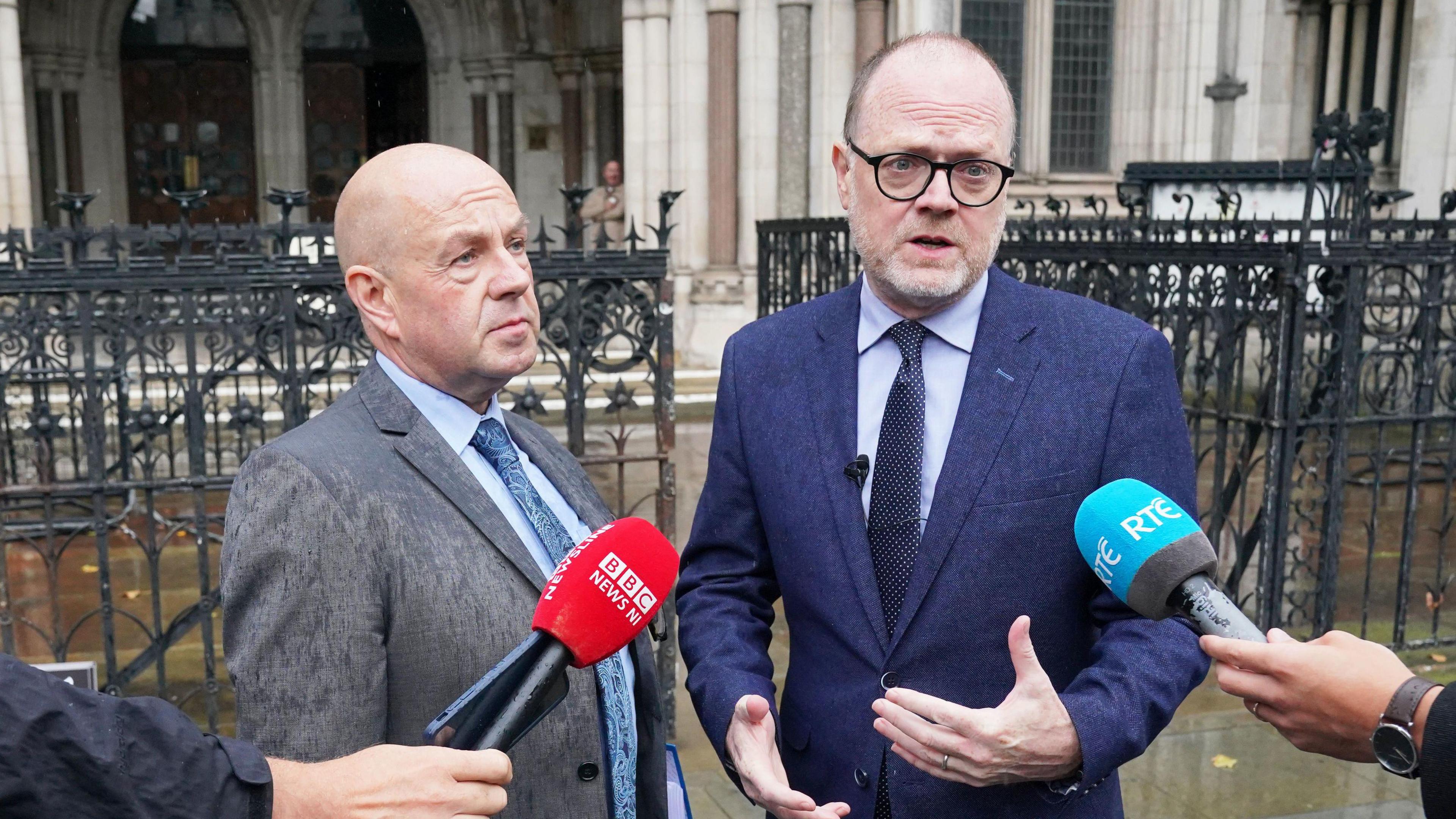 Barry on the left is bald, wearing a grey suit, blue shirt and blue tie. Trevor on the right is taller, has a bit more hair than Barry and is wearing black round glasses. He is wearing a blue shirt and dark blue navy tie under a dark navy suit. Two microphones - one a red BBC News NI and one blue RTÉ - are being held up as Trevor speaks. Behind them is the Royal Courts of Justice entrance, a grand, pillar shape with big windows as well as a black iron gate.