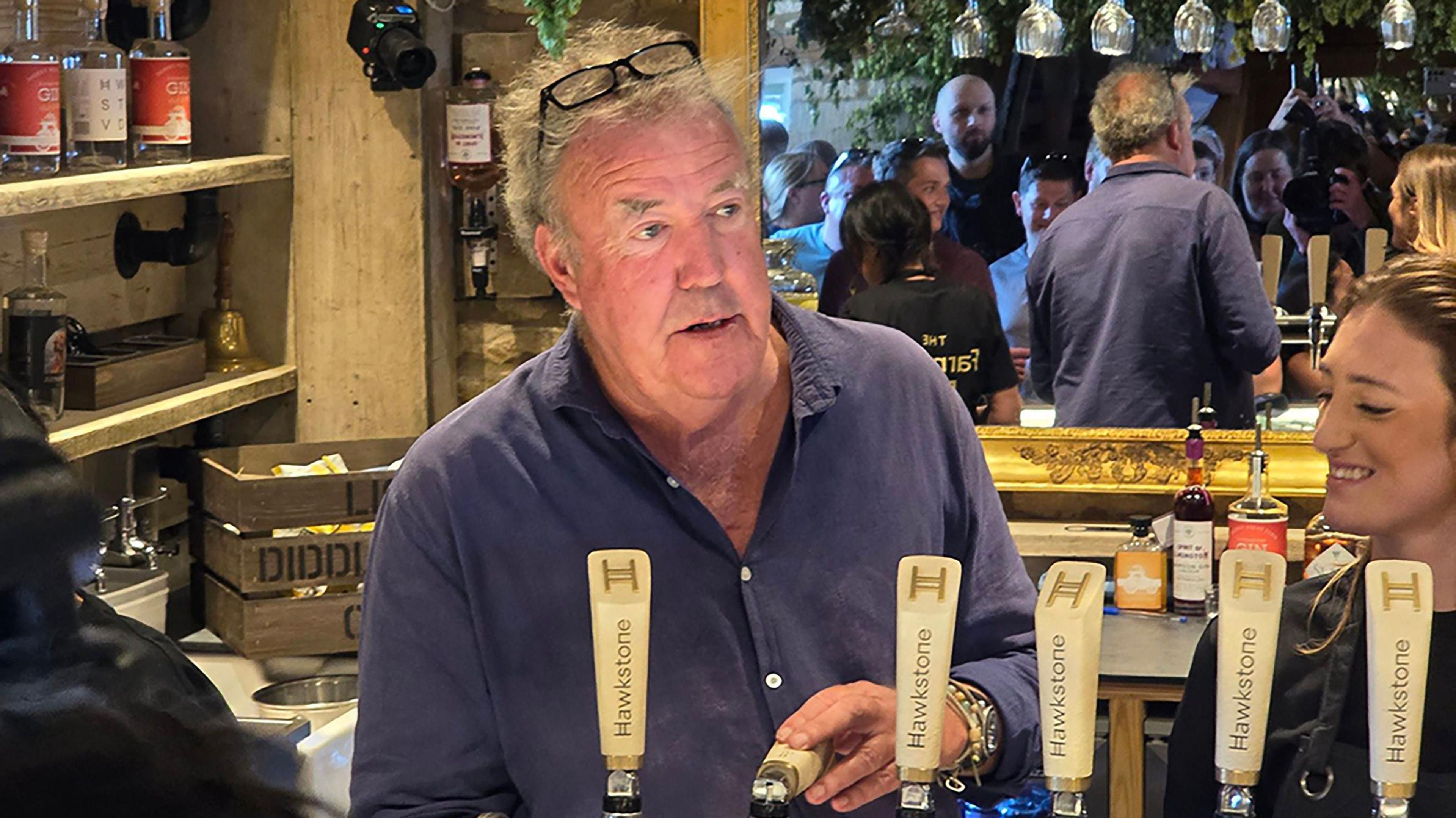 Jeremy Clarkson inside his pub, in front of a big mirror, pulling a pint. A large throng of customers can be seen in the mirror.