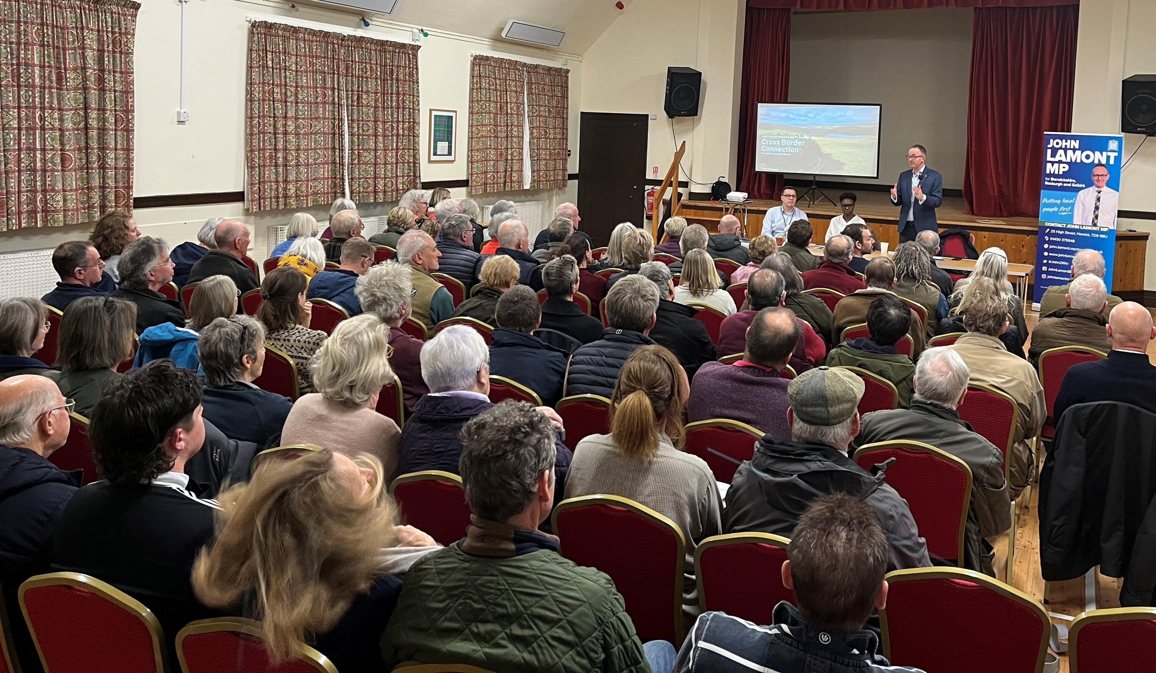 Around 100 seated people in a village hall facing a suited man who is addressing them about potential pylons. There is also a TV screen on the stage showing a landscape.