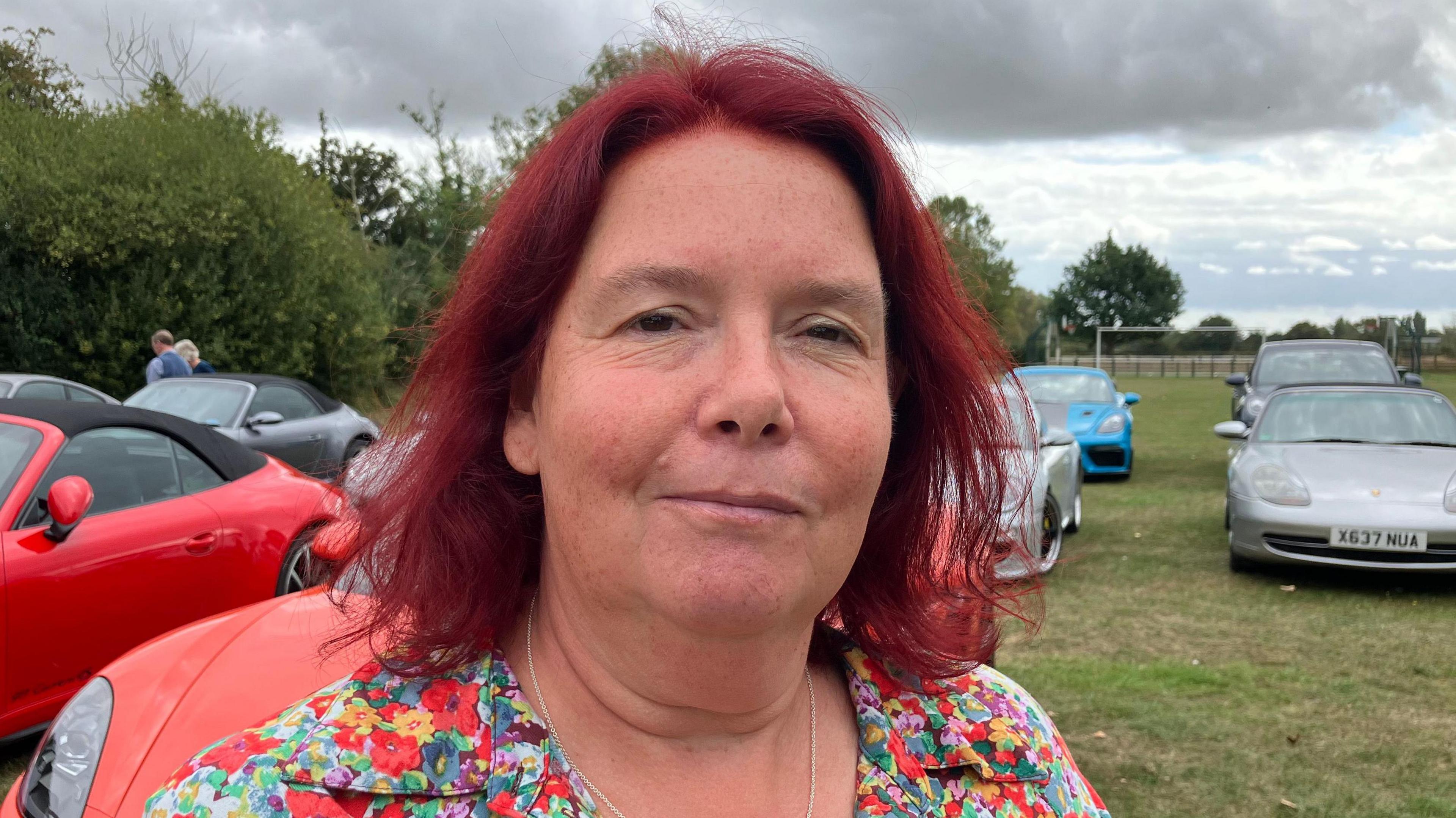 Alison Garnham is looking towards the camera with a slight smile. She has shoulder length red hair and is wearing a floral top. She is stood on a field which has rows of Porsche cars and trees.