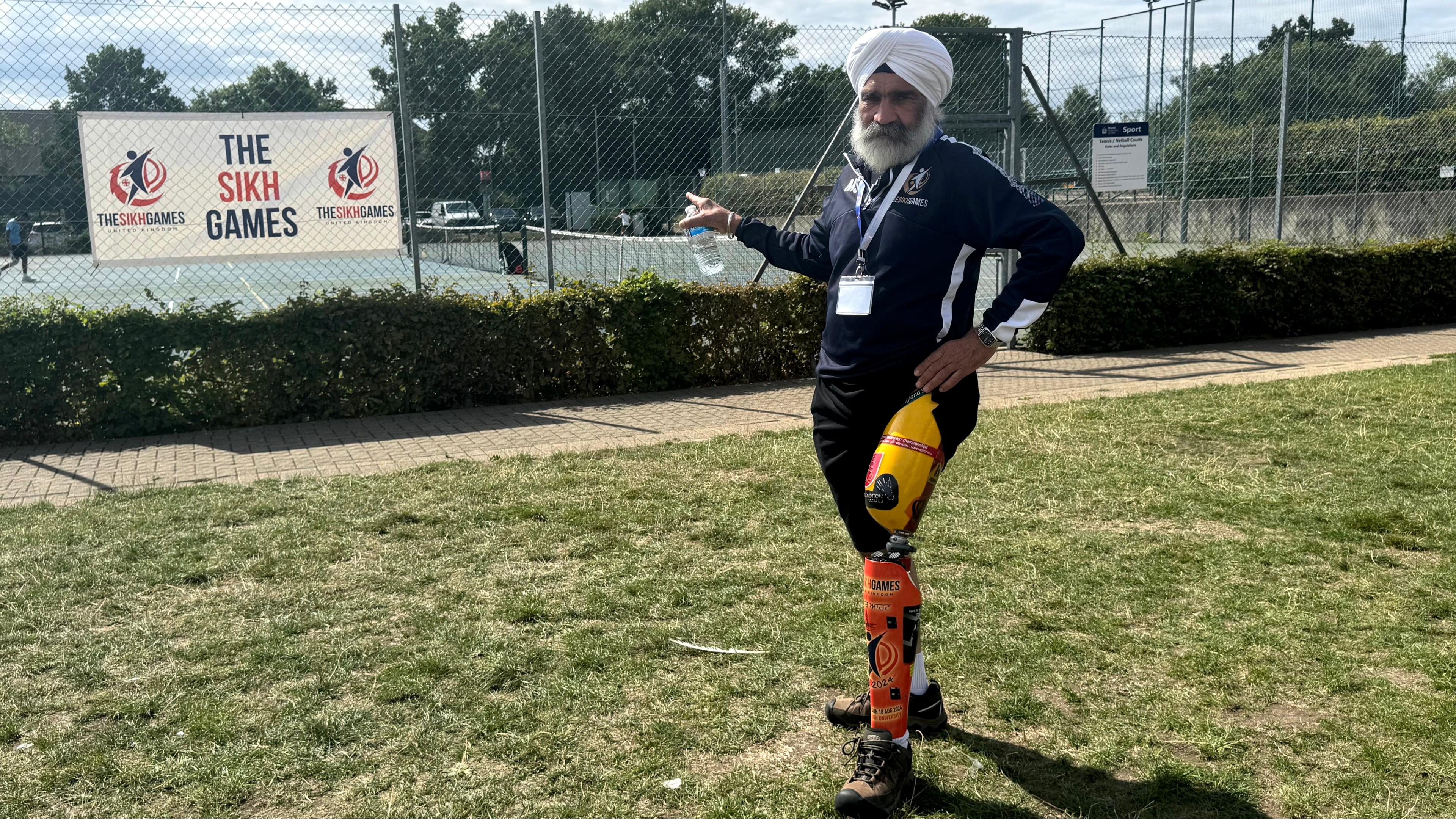 Meva has a white turban and white beard. His left leg is prosthetic. He wears black jogging bottoms and a dark blue quarter-zip sweater while pointing to a sign saying the Sikh games. 