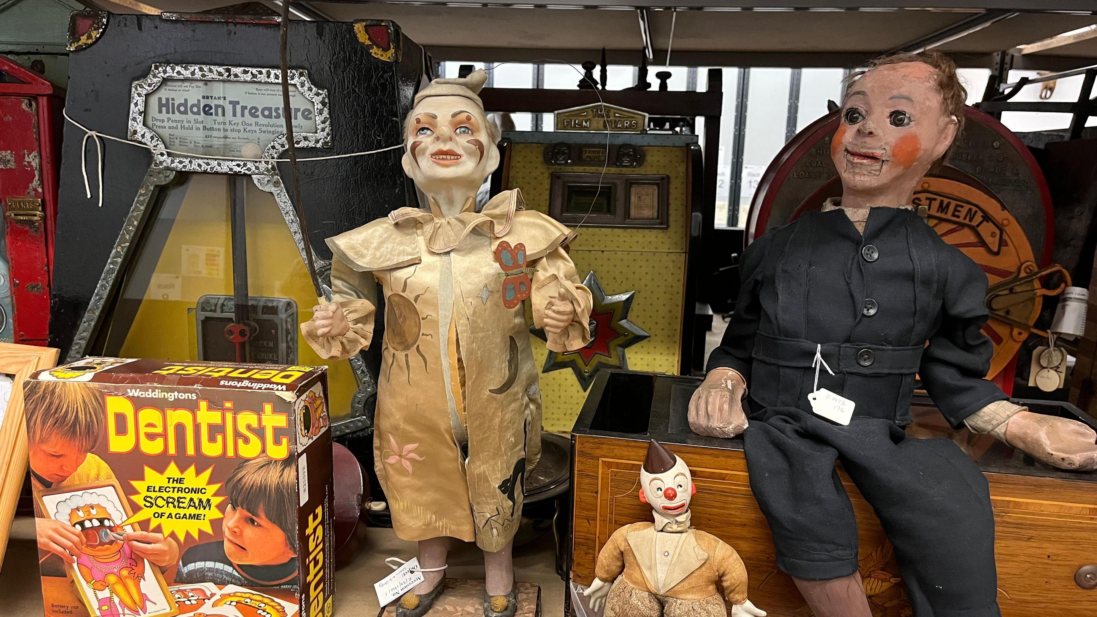 A shelf displaying vintage and antique items, including two old-fashioned dolls dressed in worn clothing. One in a beige coat with decorative patches and the other in a dark suit. In front of them is a small clown doll with a painted face and fabric body. To the left, there is a brightly coloured retro board game box titled “Dentist: The Electronic Scream Game,” featuring illustrations of children playing.