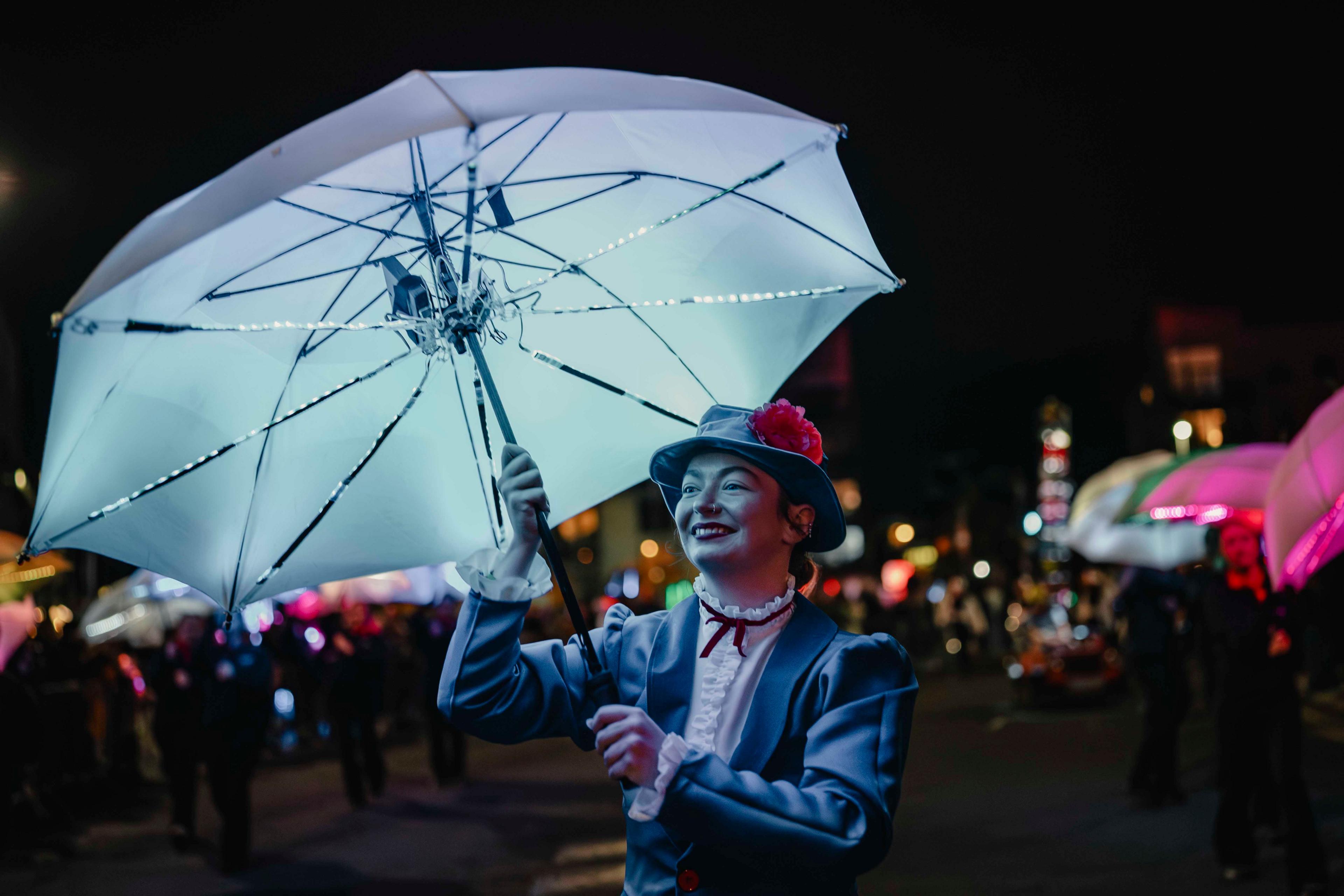 A woman dressed as Mary Poppins holding a large white umbrella which is illuminated with LED lights. She smiles and walks as part of a glowing parade against a black night sky.