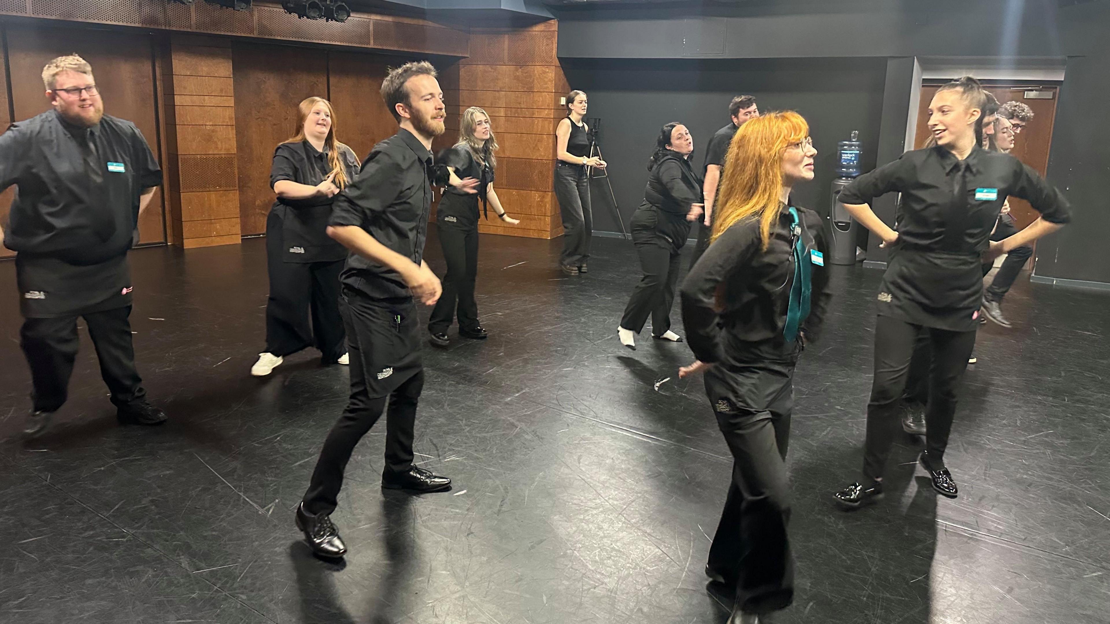 People all in black rehearsing a dance in a room with shiny black floor and brown brick walls.