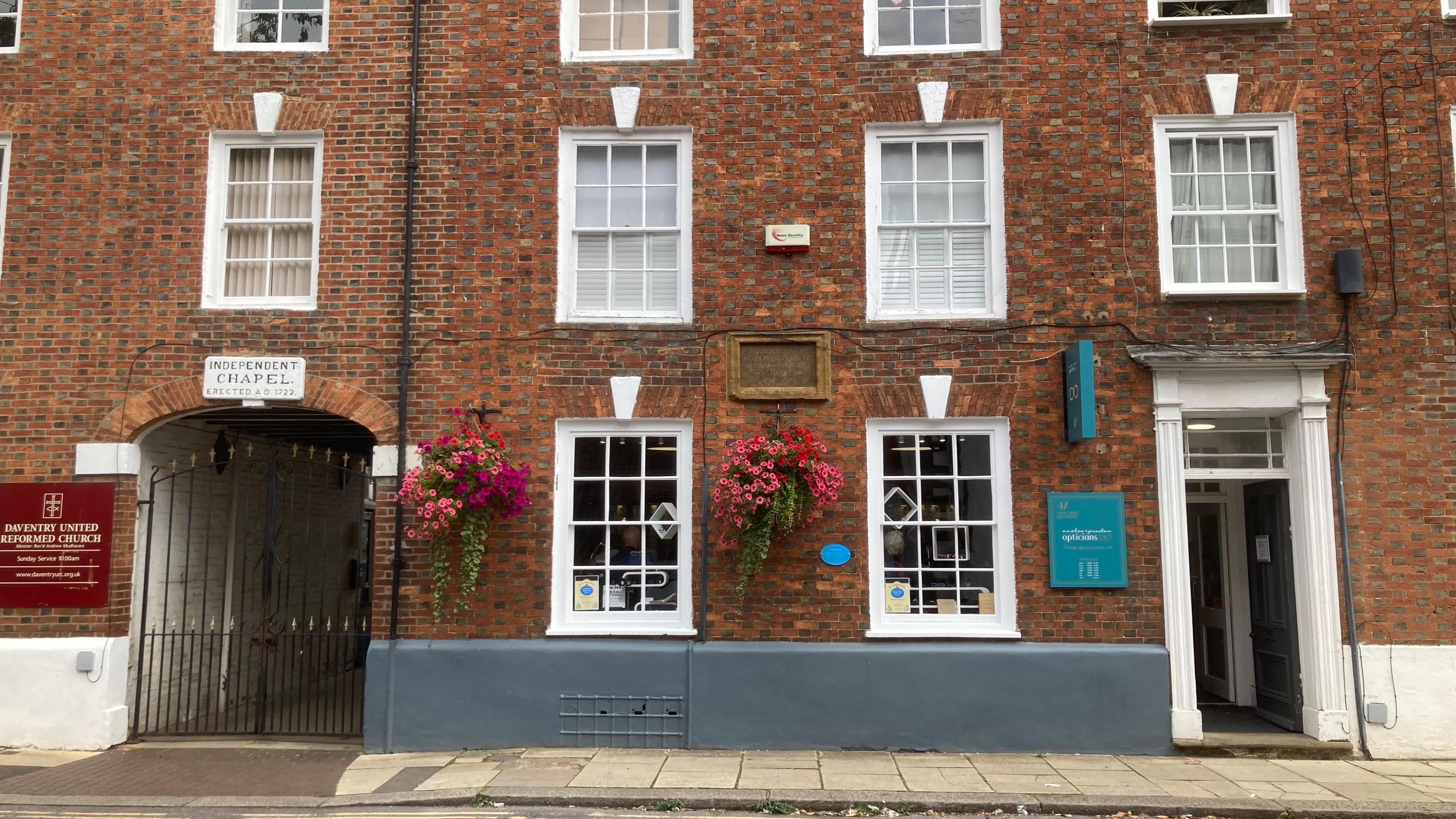 Brick building with high windows alongside black railed gate with adjacent sign reading "Daventry United Reformed Church"