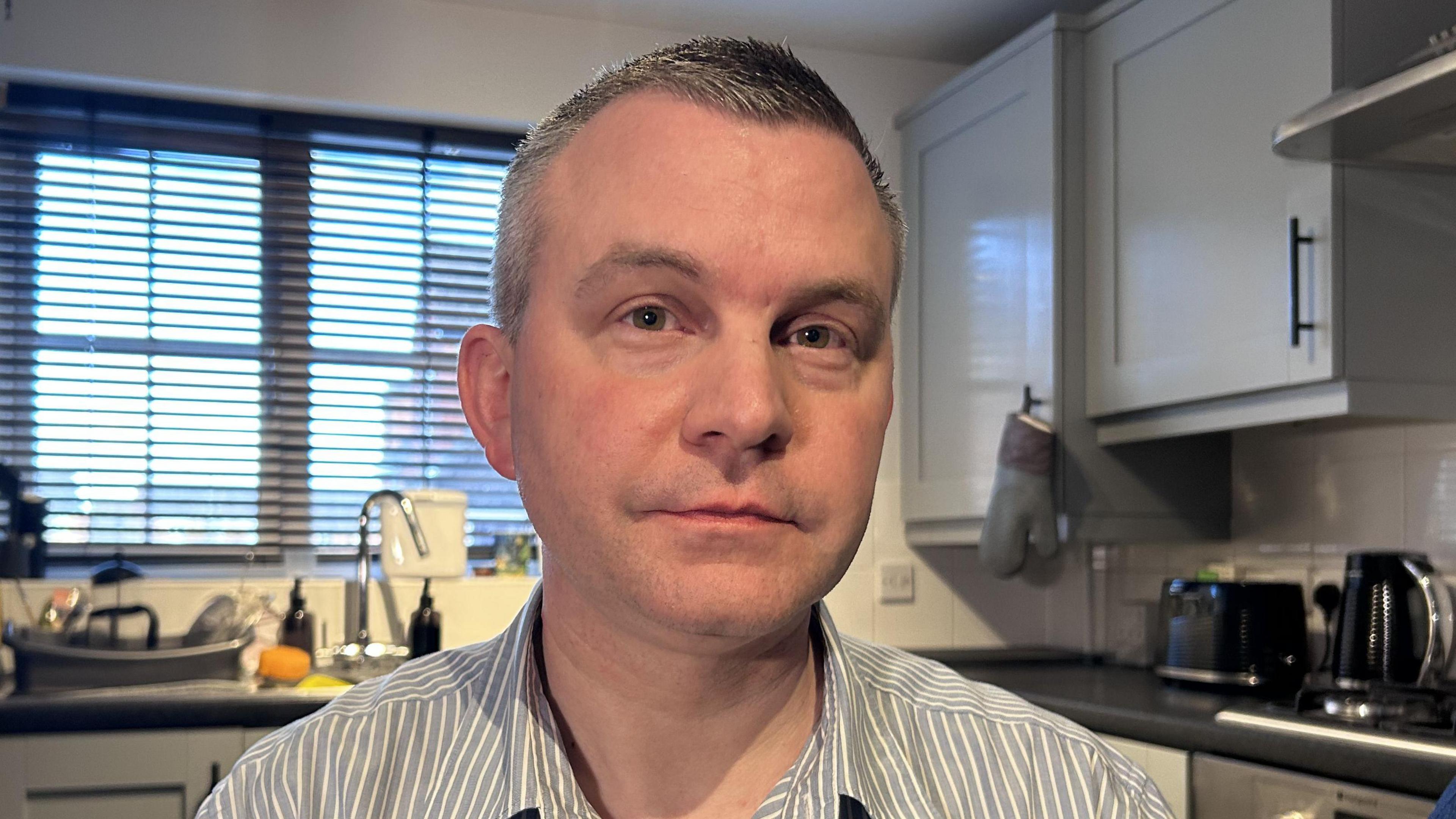 Stephen with short hair sits in a kitchen. He is wearing a grey and white striped shirt.