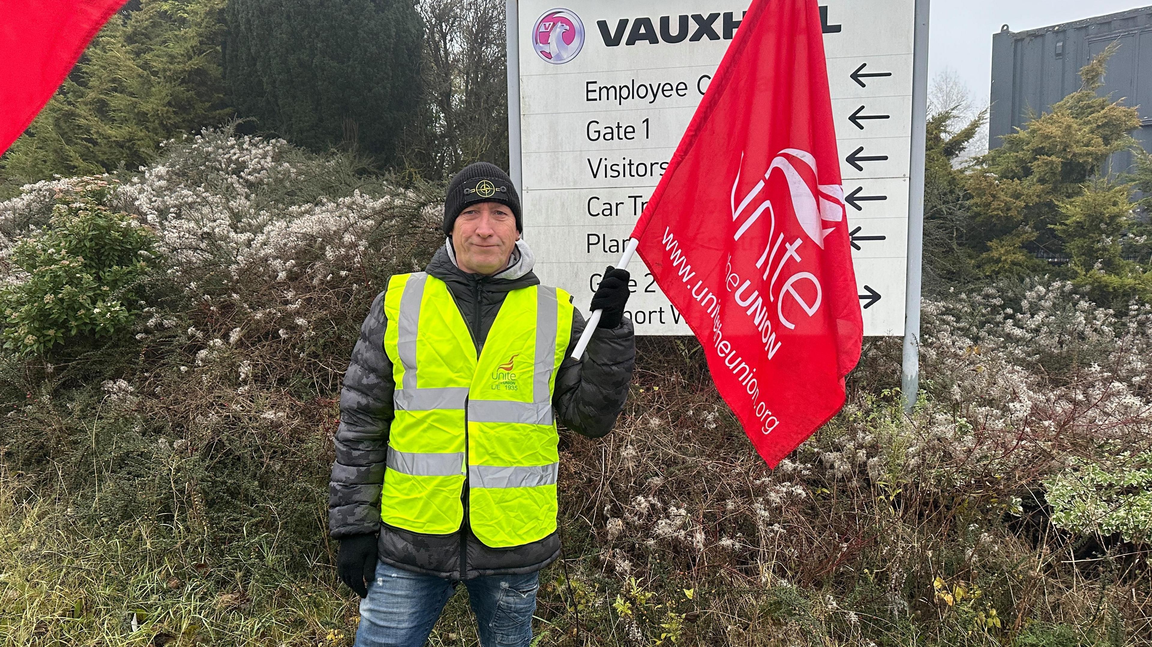 Paul Geary wearing a high-vis jacket over his black coat and wearing a black hat. He is waving a red Unite flag outside Vauxhall in Luton