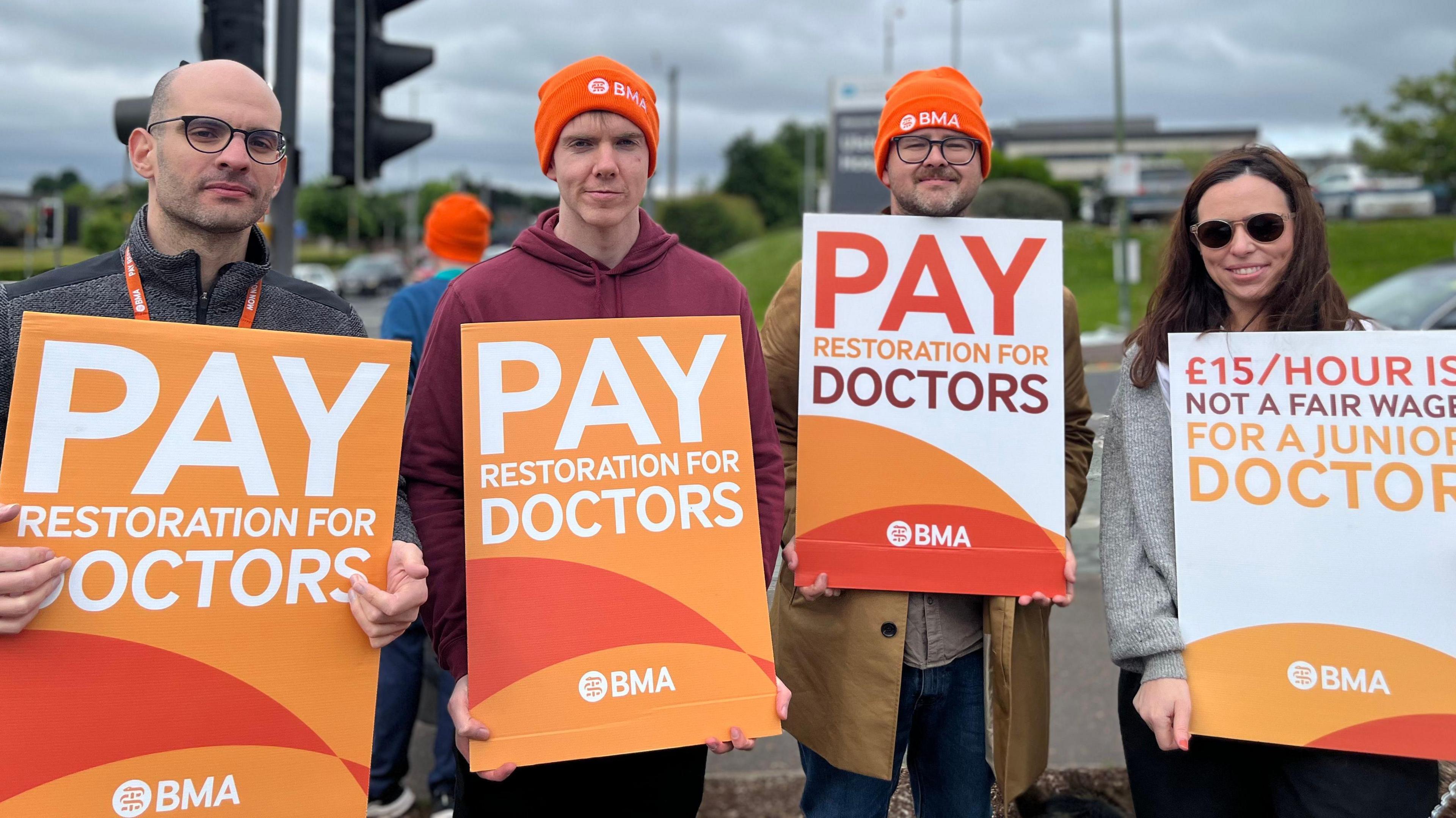 Four people - three men and a woman - are holding orange and white signs outside a hospital. On three of the signs it reads 'pay restoration for doctors', and the fourth sign reads '£15/hour is not a fair wage for a junior doctor'.