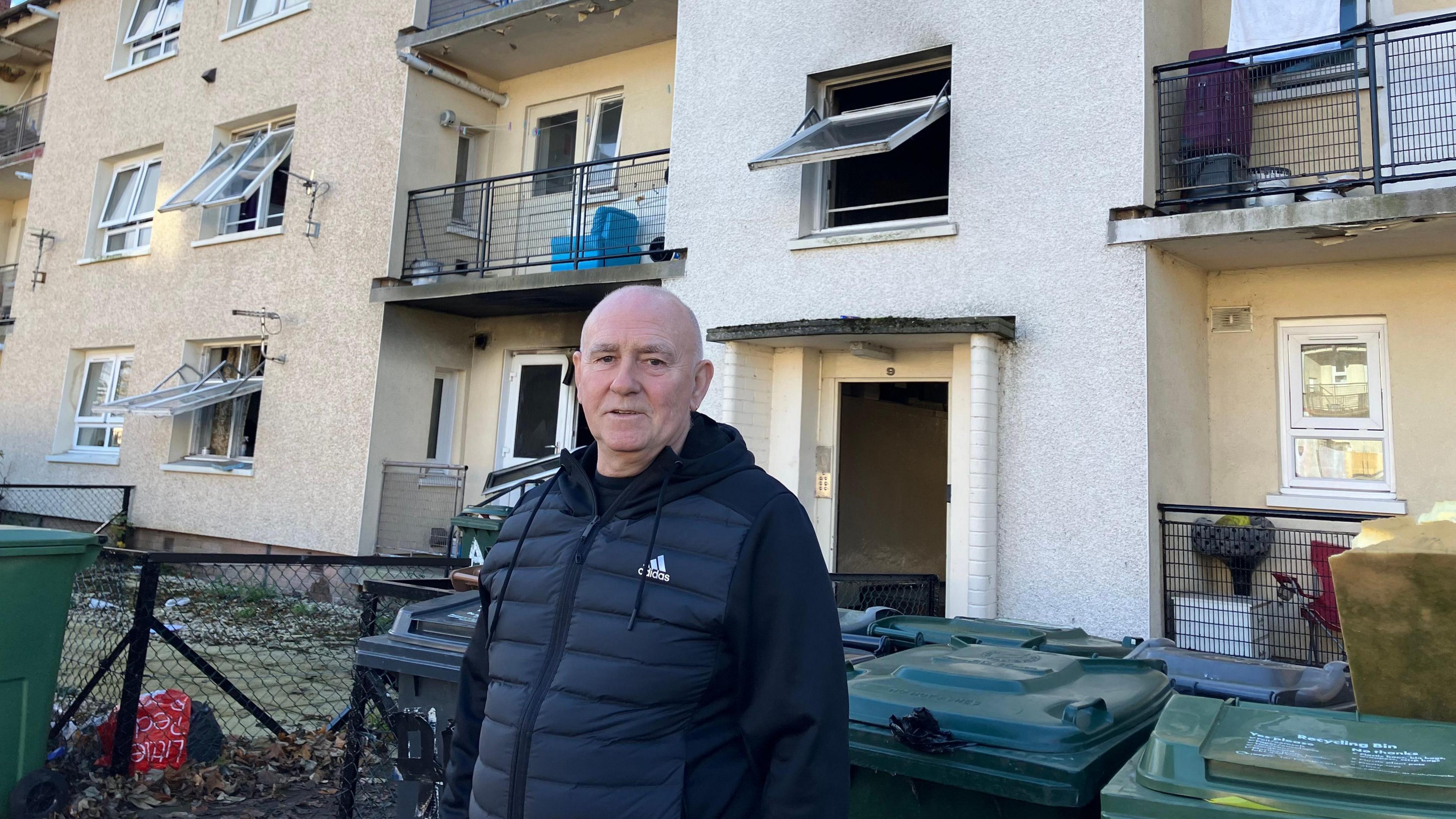 John Hill standing in front of the block of flats in Niddrie Mill Crescent. John is bald, middle aged and is wearing a black hooded jumper with a zip and silver Adidas logo. Behind him is the block of flats, which is built from light stone. There are visible signs of black charring to the brick work.