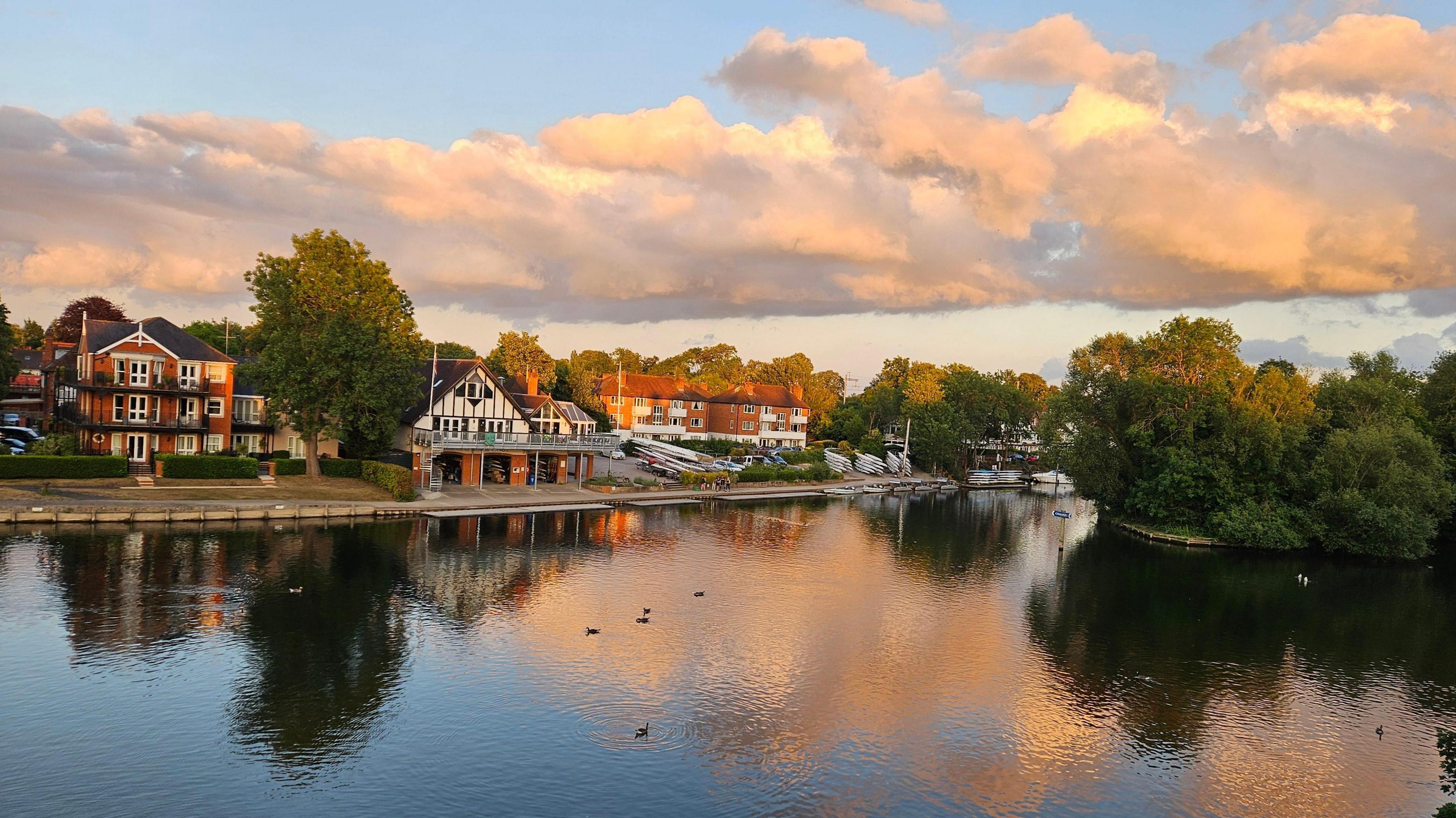 The River Thames reflects the fluffy clouds and blue and yellow skies next to a boathouse and homes on it's banks. A few birds can be seen floating on the water.