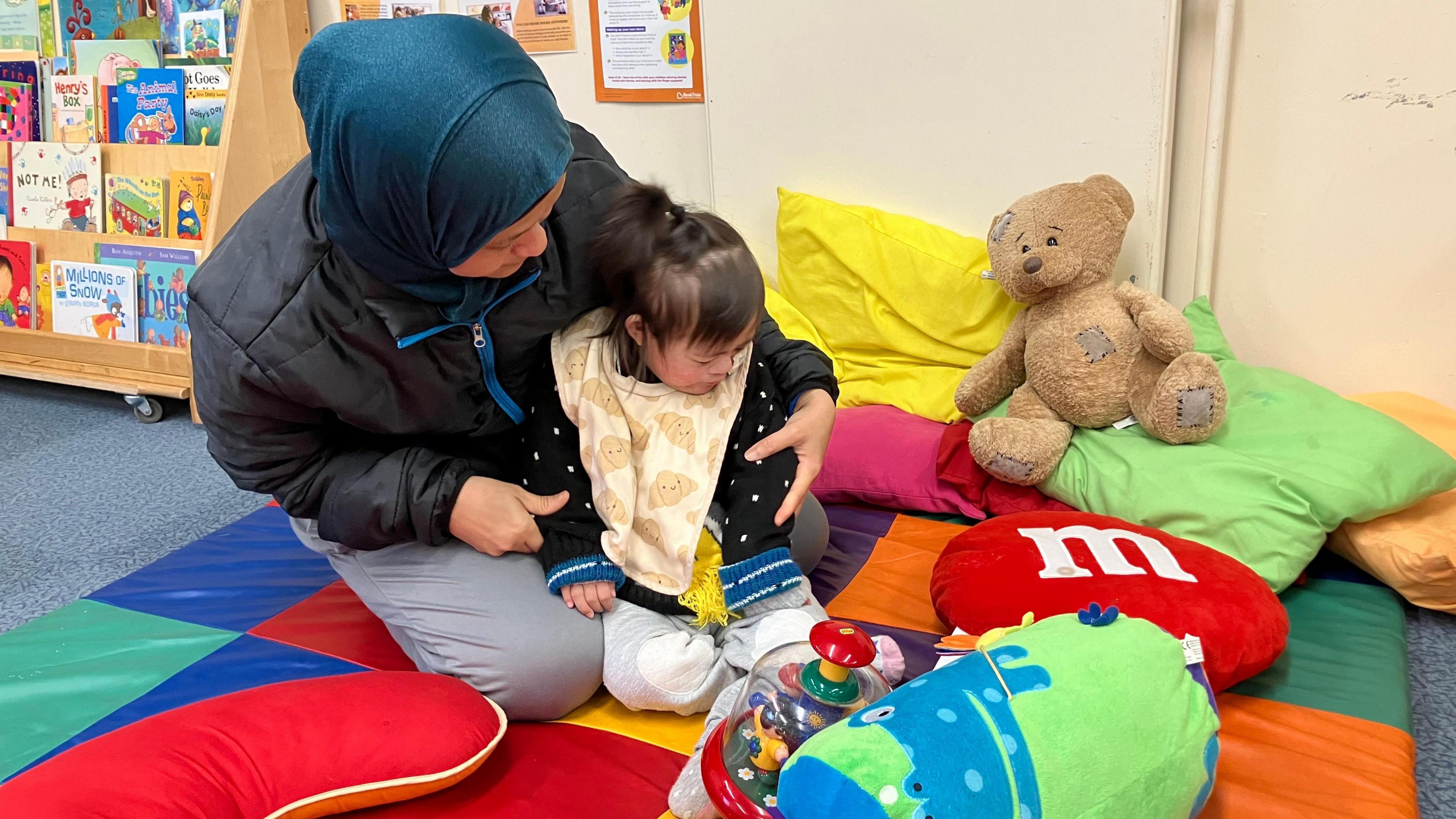 Leni wearing a blue head-scarf, a black puffer coat and grey trousers- witting on a colourful play mat holding her daughter- wearing a black cardigan and a yellow bib