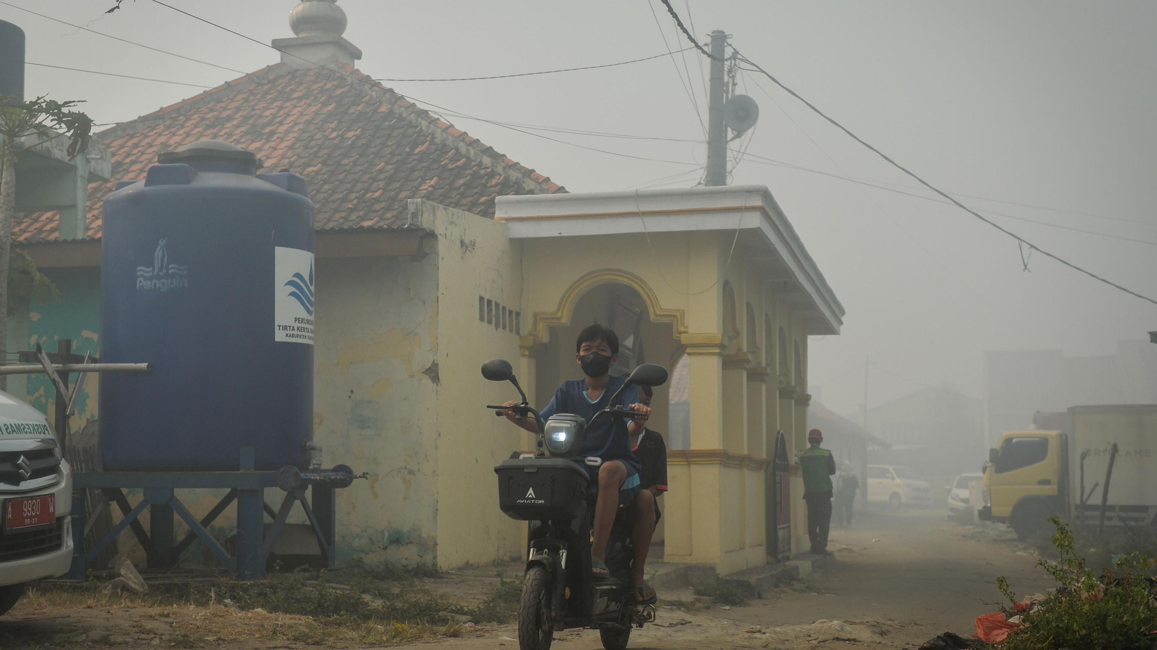 A person wearing a face mask drives a motorbike down a road beside a house, while the air is shrouded in smoke