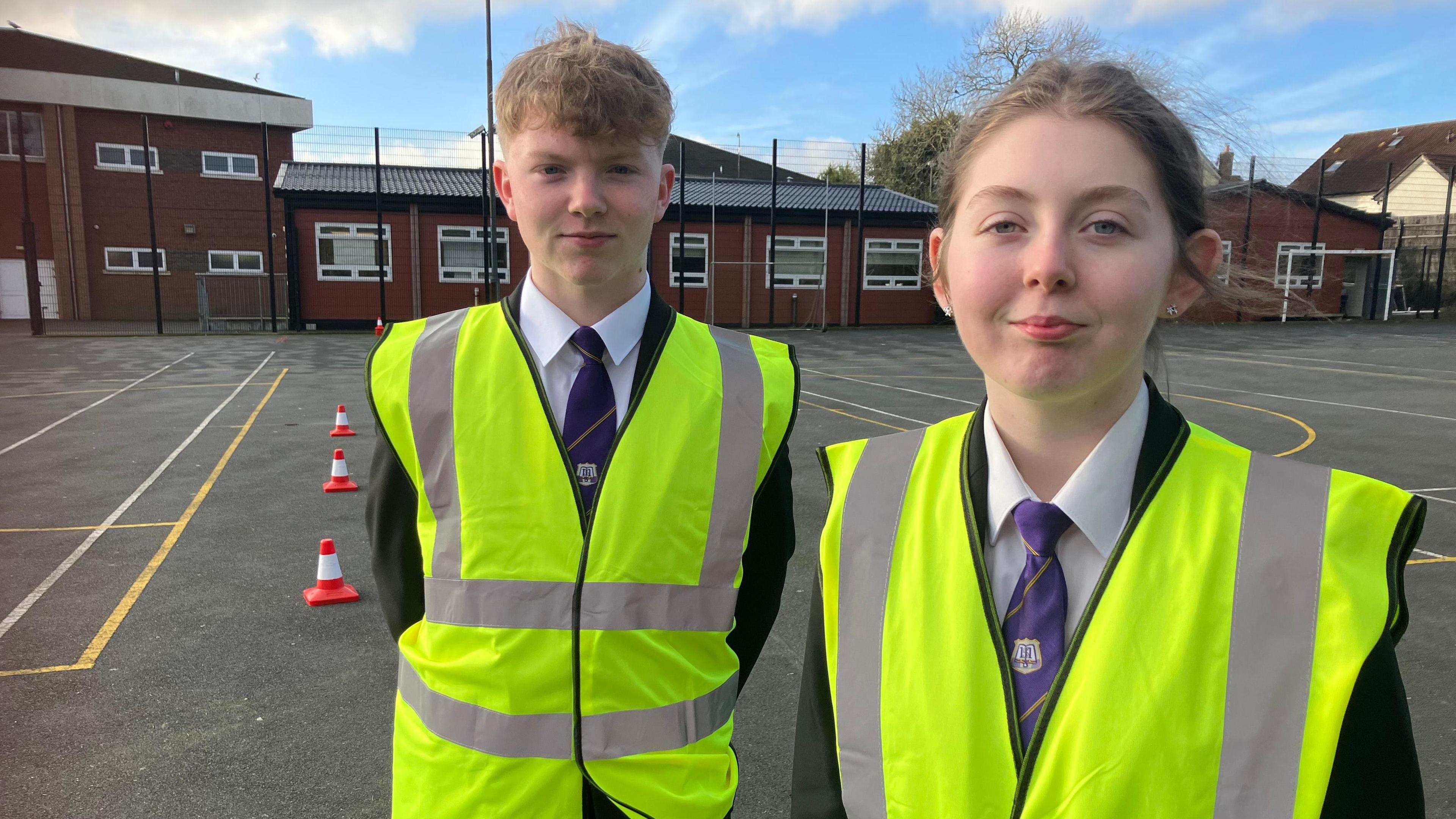 Matthew Blair (left) and Amy McCoy (right). Matthew has strawberry blonde hair, Amy has brown hair tied in a ponytail. Both are wearing their school uniform - black blazer, white shirt and blue tie - and high viz vests. They are in the school playground with the school building in the background.