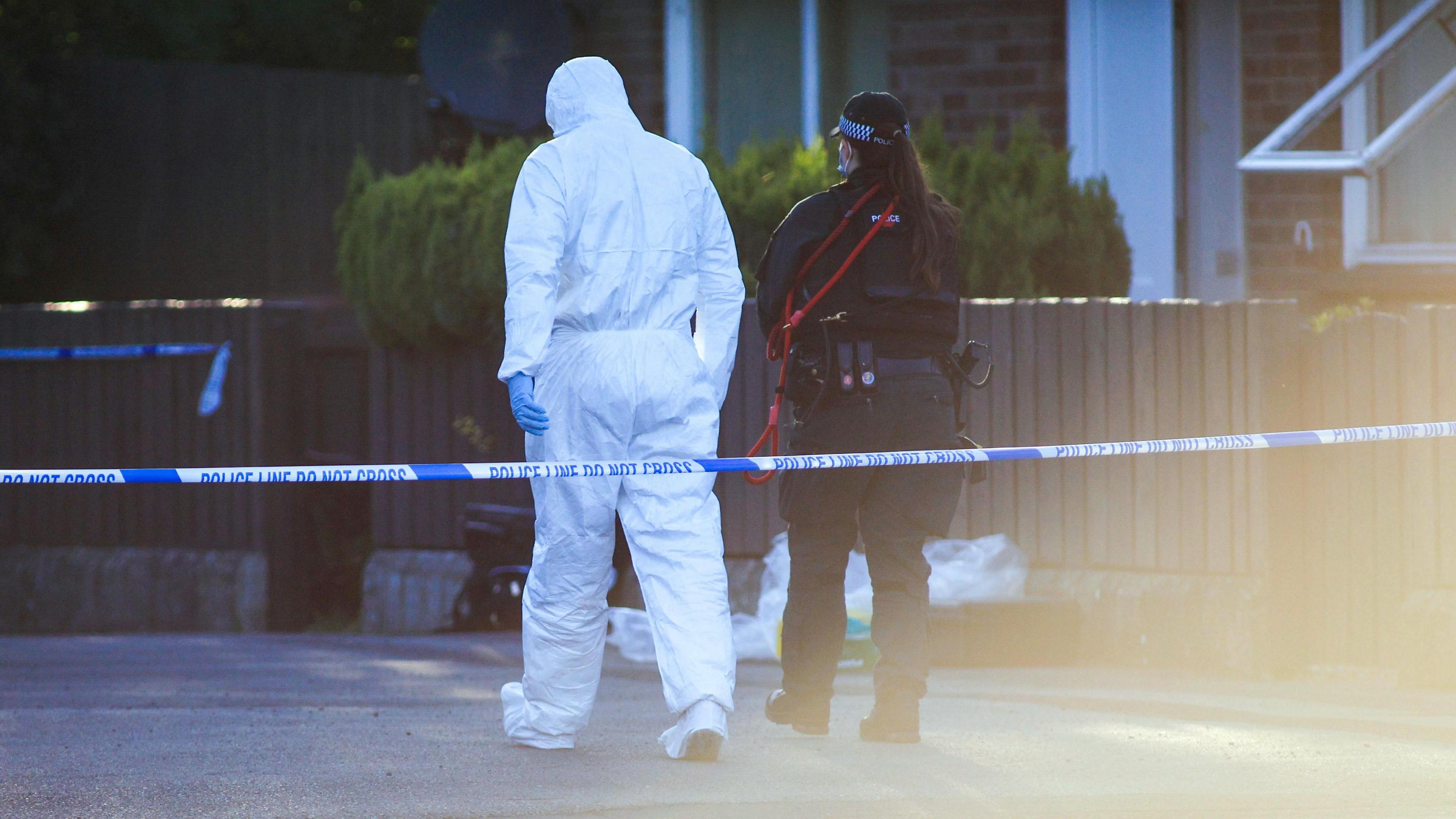 Forensic officer in white suit and police woman in dark uniform with their backs to camera, stood outside a row of bungalows with a low wooden fence, there is police tape cordoning off the road