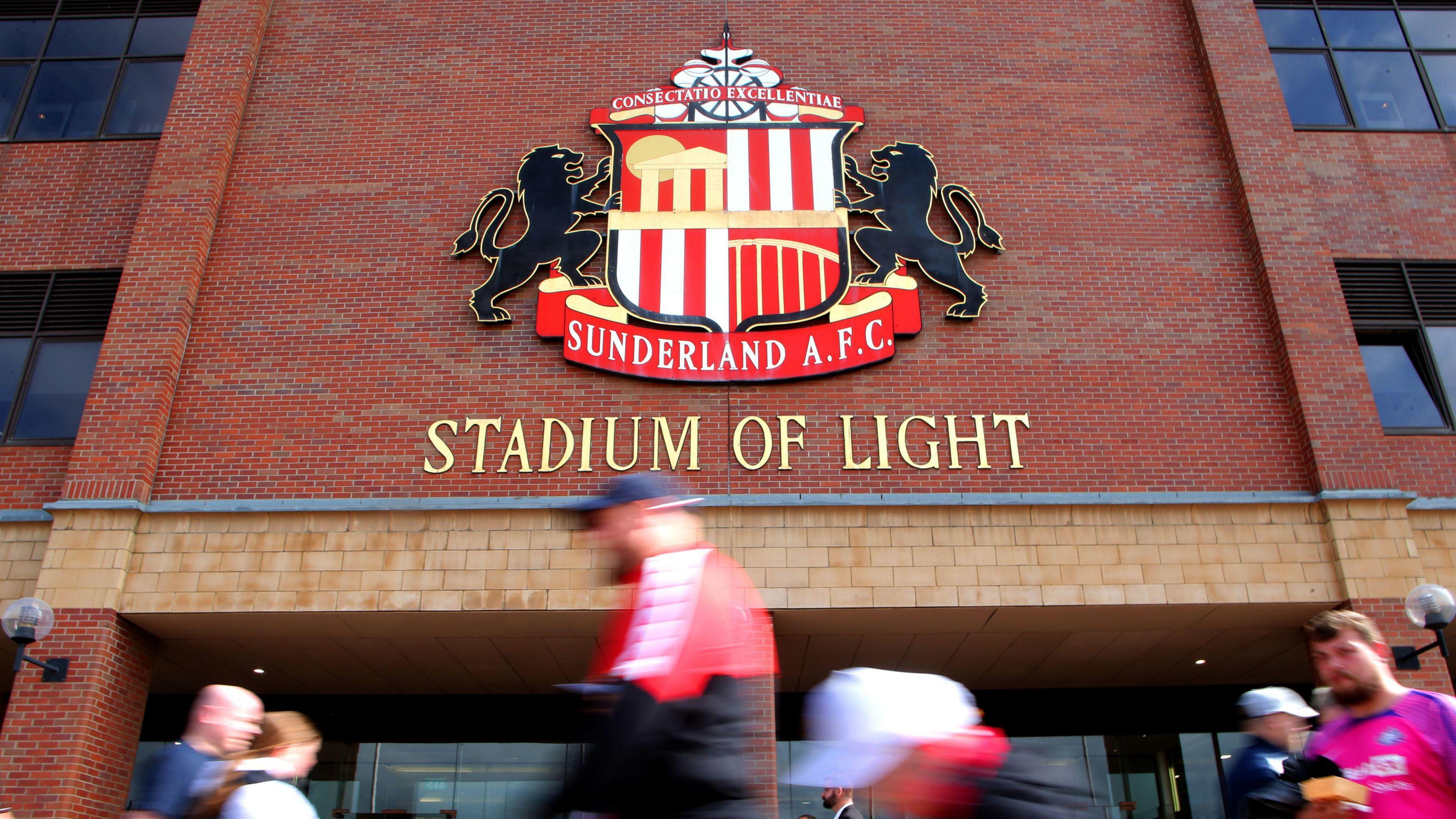 A general view of the Stadium of Light, Sunderland. The Sunderland logo is mounted on a brick wall, with people walking beneath the sign.