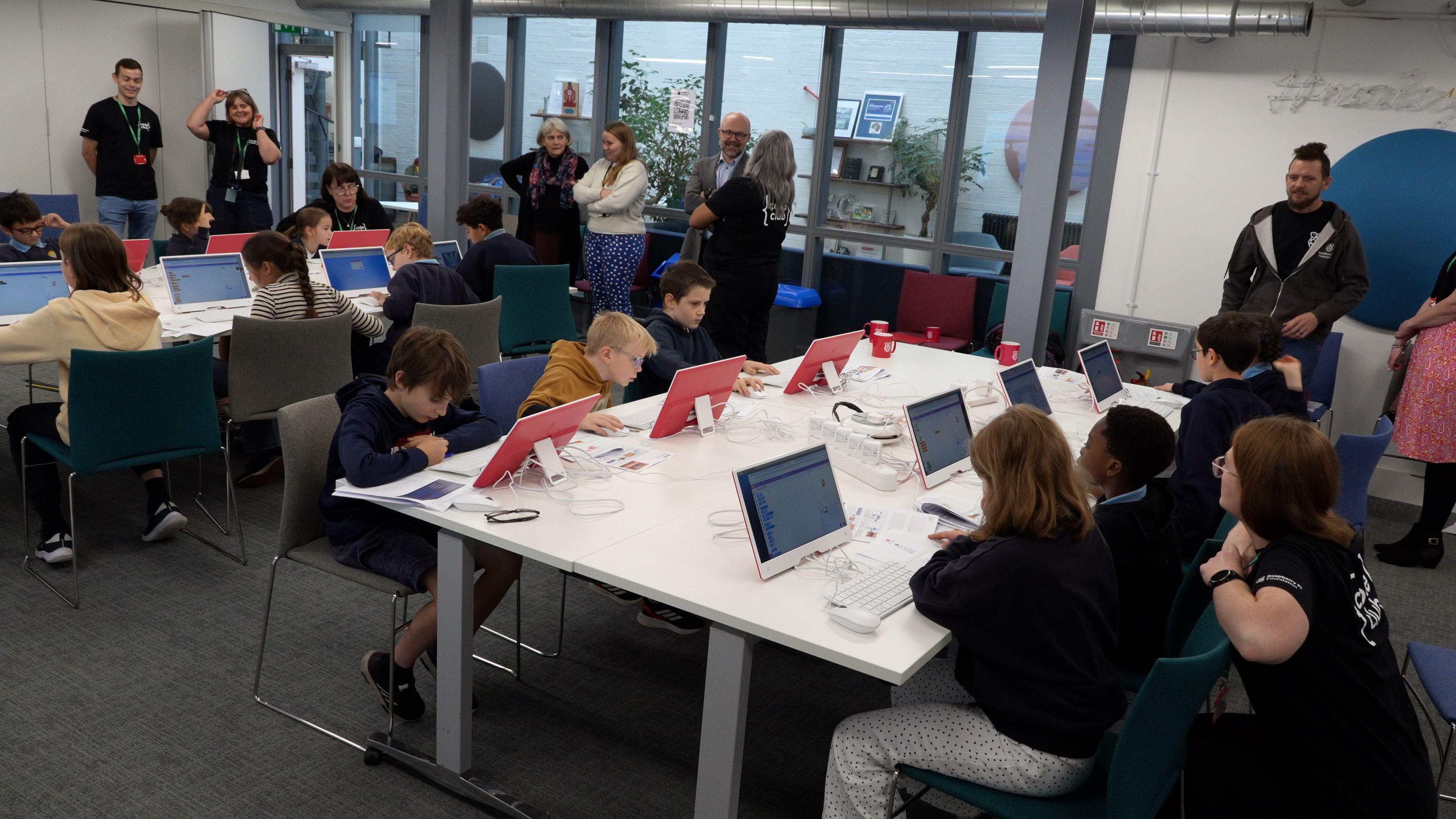 A group of children sitting around a large table, each focused on their own laptop as they take part in a tech-based workshop. Several adults stand nearby, offering guidance and support.
