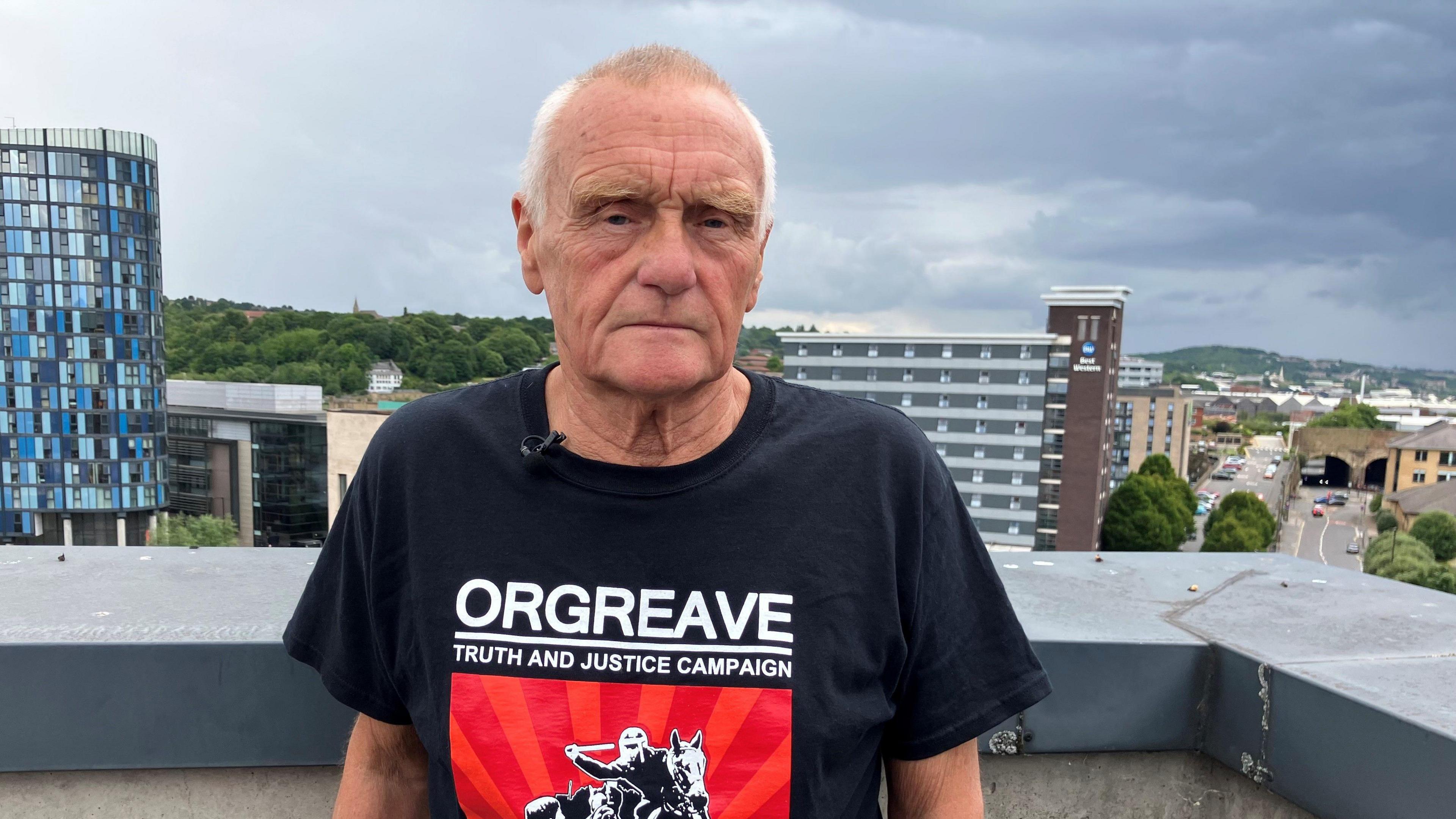 John Dunn has short grey hair and looks towards the camera he is wearing a black and red T-shirt wiht the words Orgreave Truth and Justice Campaign written on it