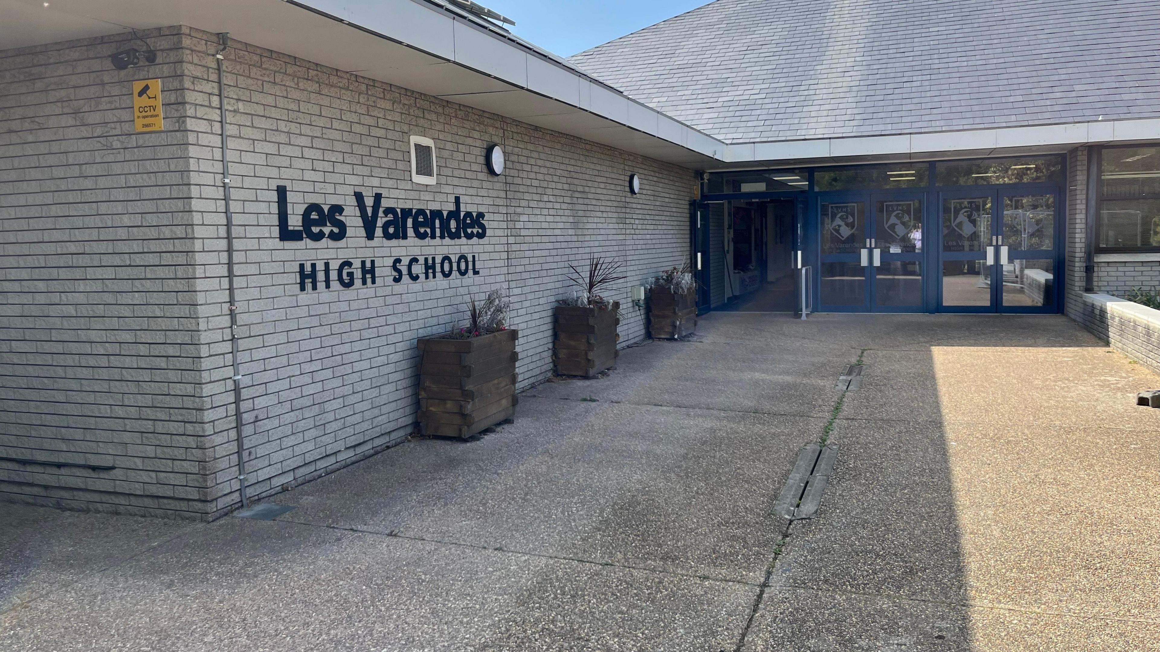 An exterior picture of Les Varendes High School, there is a large sign with the name. The building is a grey brick building with blue rimmed glass doors. 