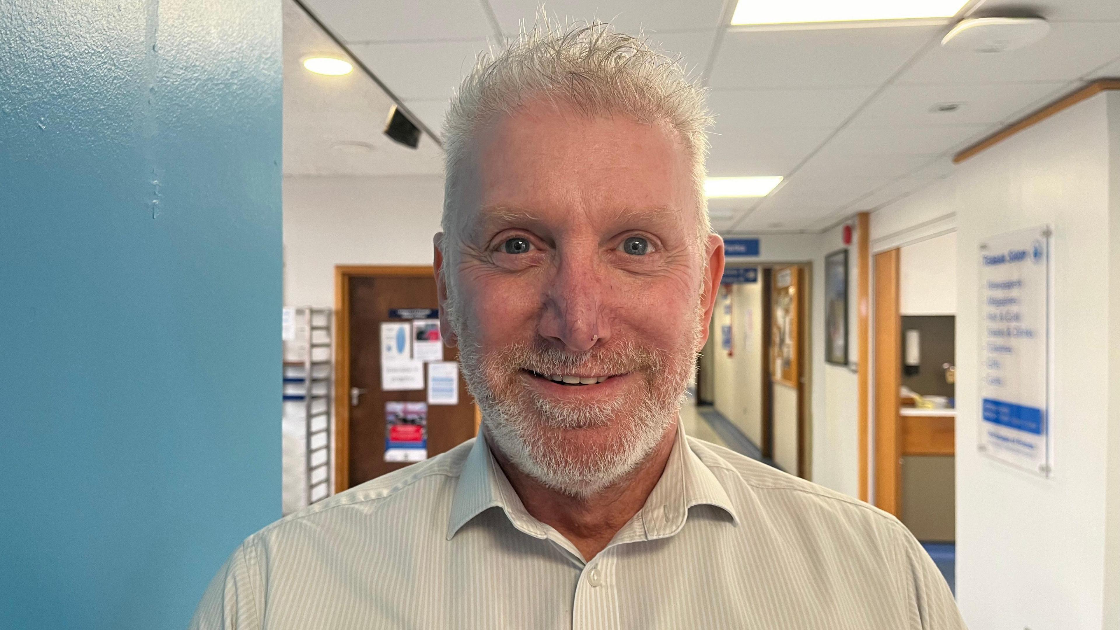 Ali looking into the camera and smiling; he's wearing a white collared shirt. Behind him you can see a hospital corridor with blue and white walls. To his right you can see signs directing visitors and patients around the hospital, but they're not in focus so you can't see what they say. 