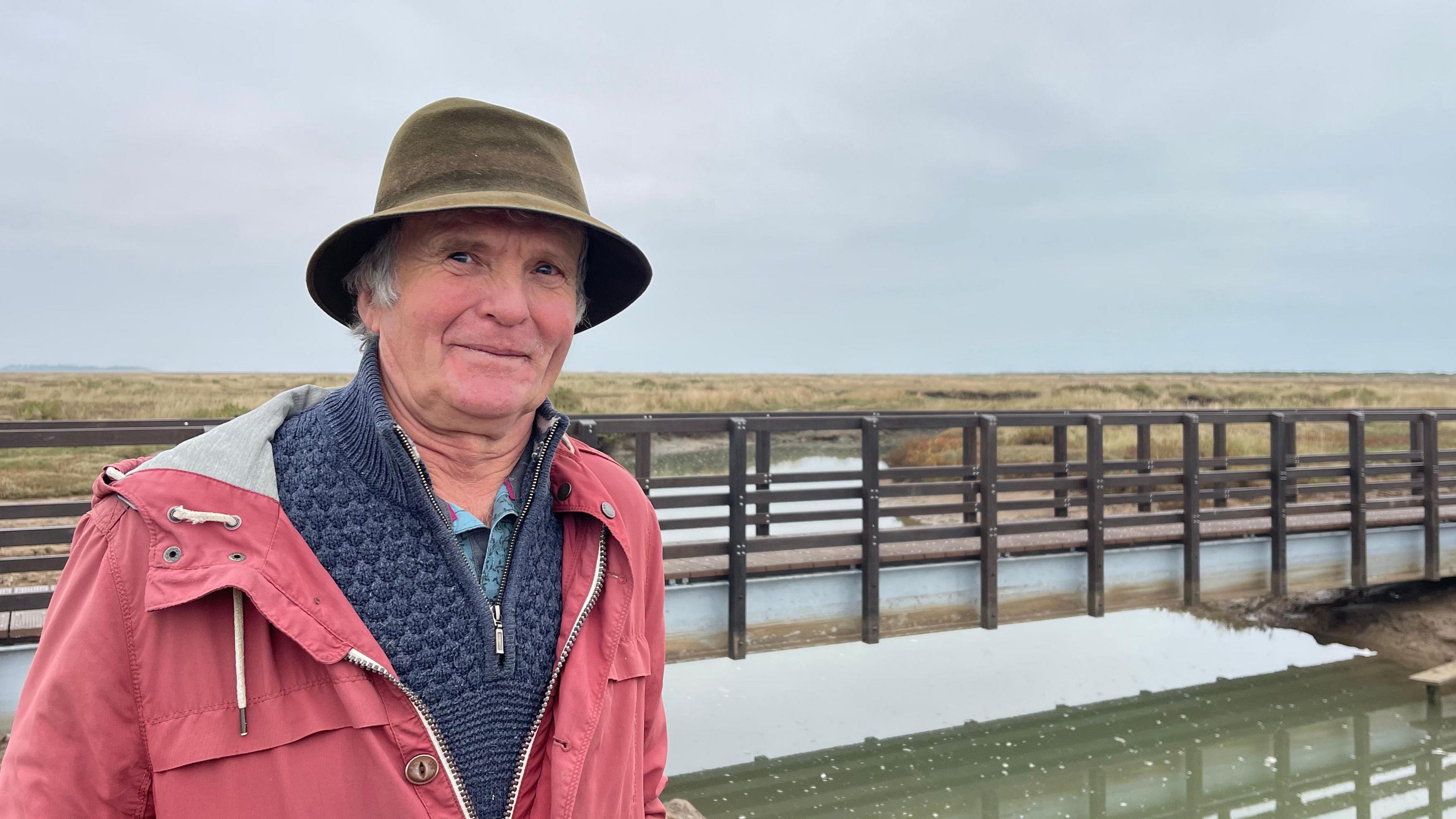 Ian Curtis wearing a khaki-coloured hat, a blue jumper and red raincoat. He is standing in front of the new Stiffkey bridge.