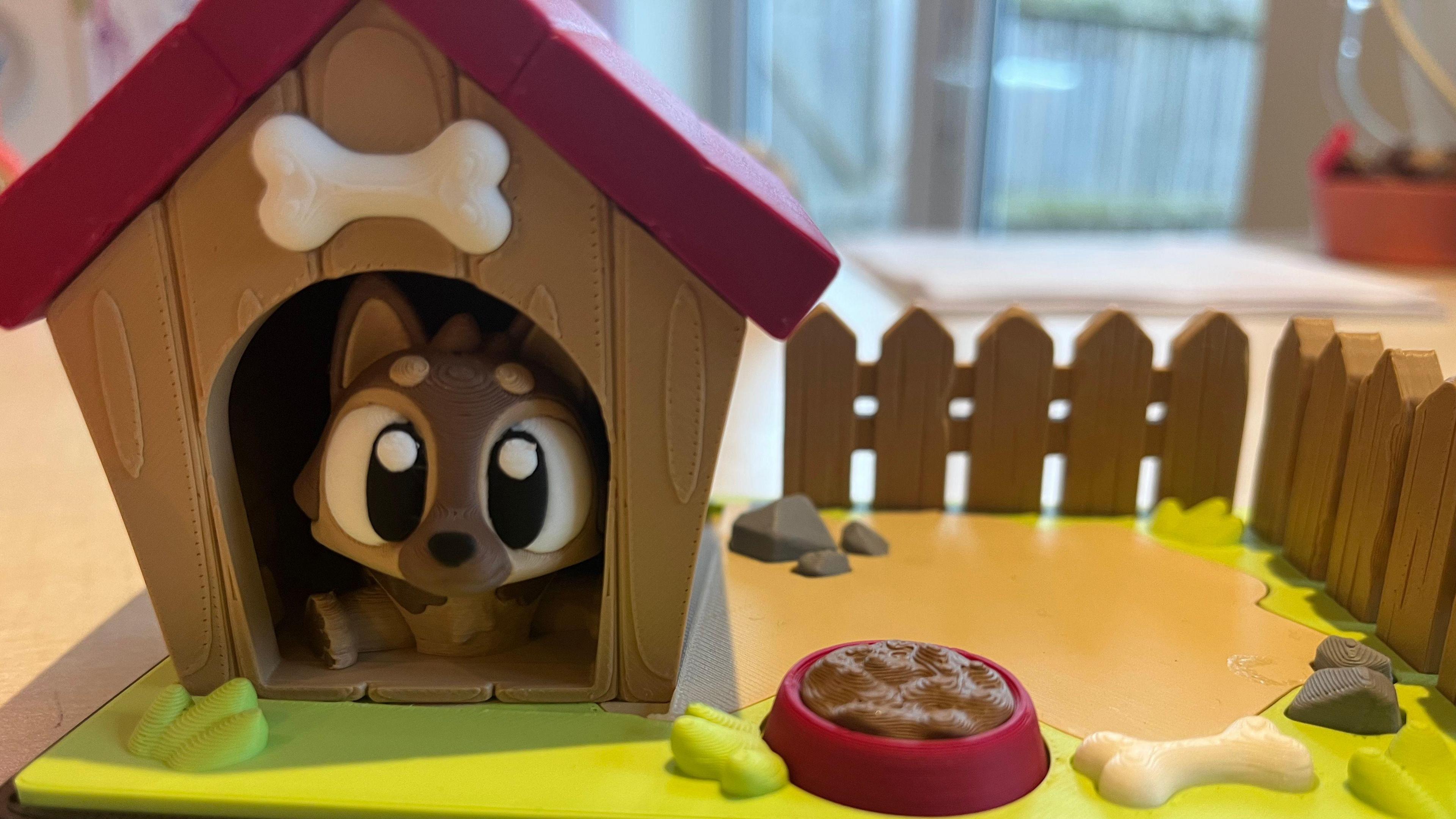 A 3D printed toy of a cartoon-like brown dog with big eyes sitting in a kennel with a red roof with a bone above the entrance. The kennel is sitting in a fenced area with a bone and a bowl of dog food.