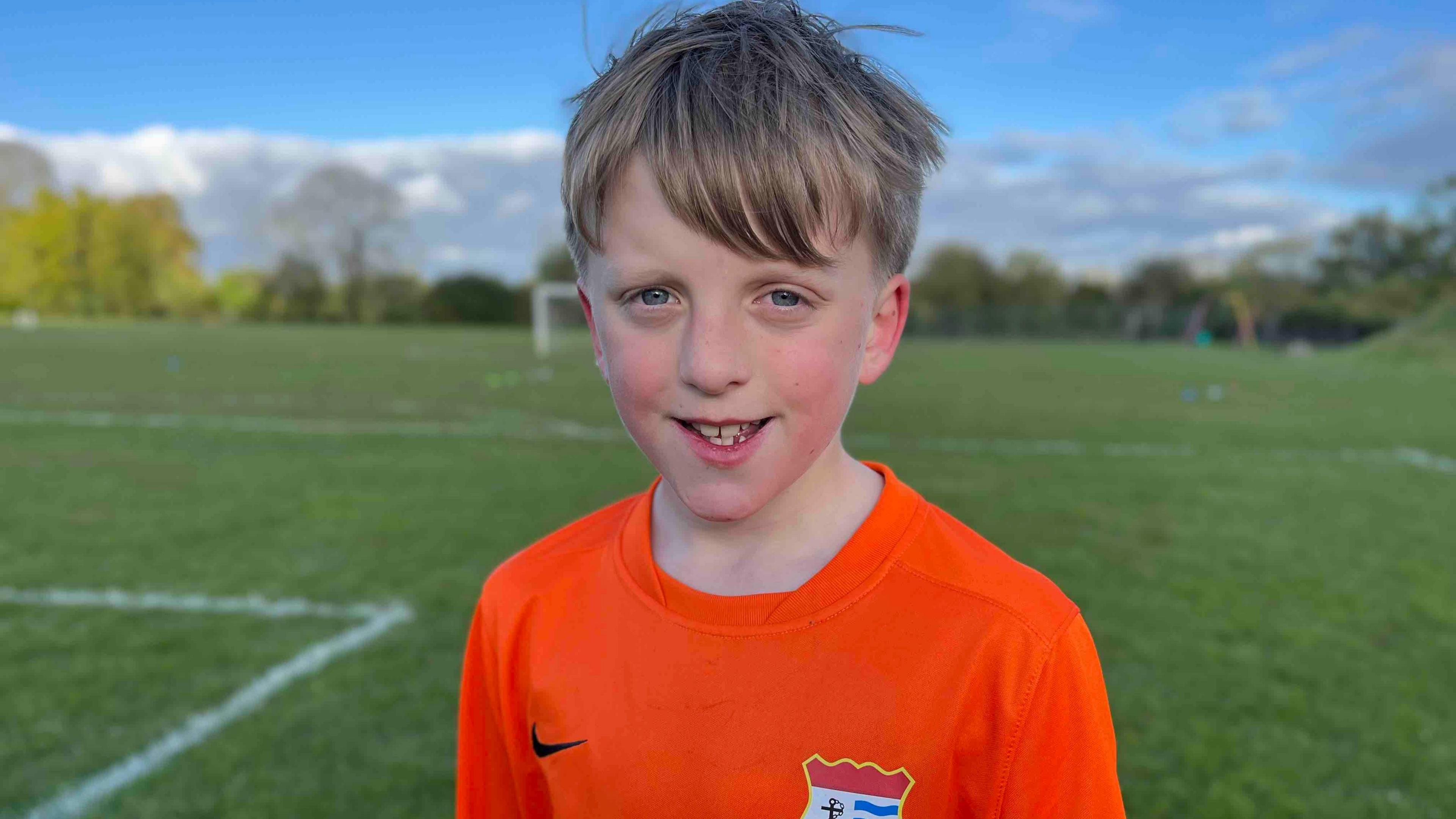 Harry standing in an orange football shirt, with a pitch behind him