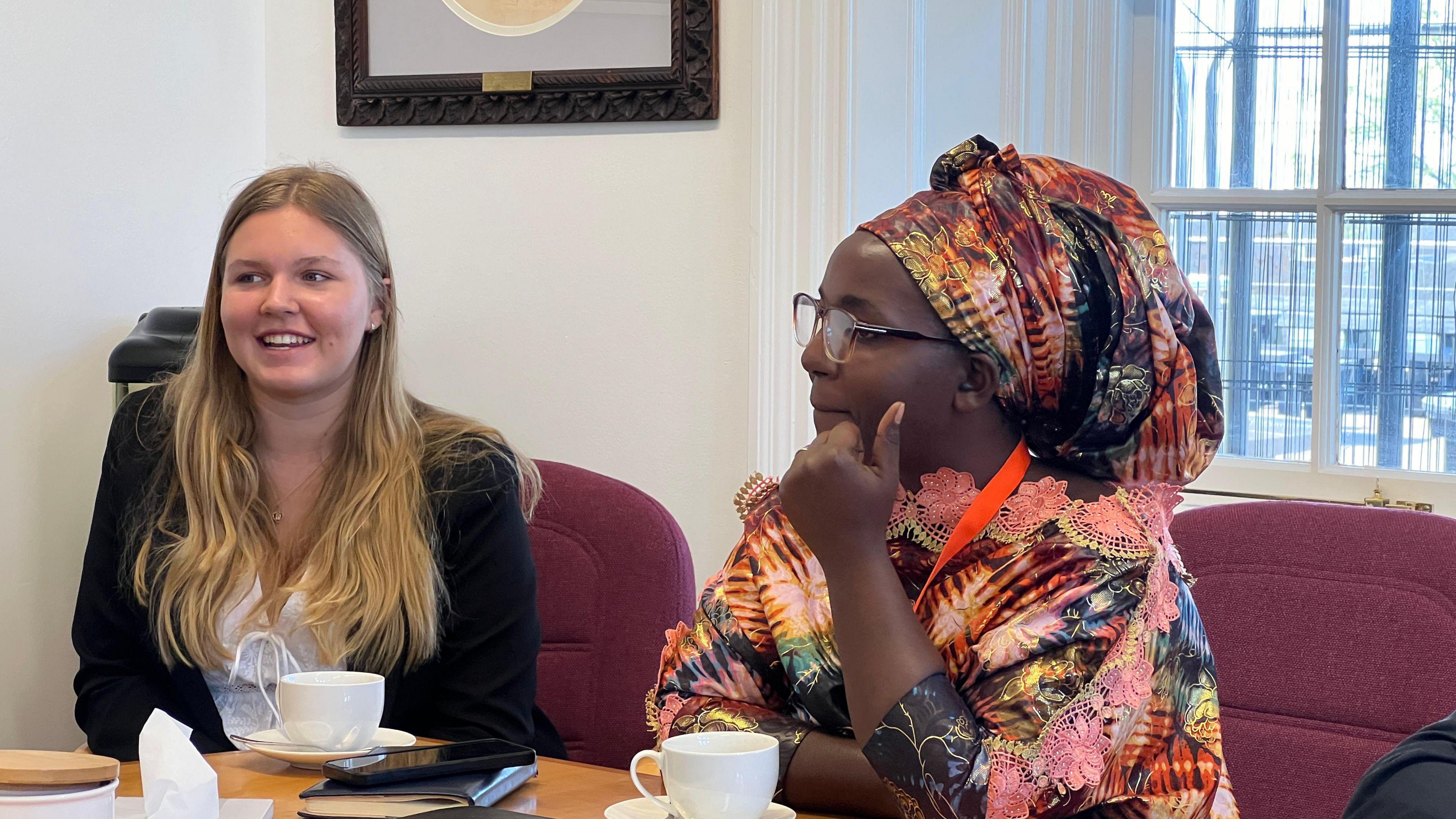 Yasmine smiles as a head teacher in traditional African dress sits next to hear as they both have cups of tea in front of them