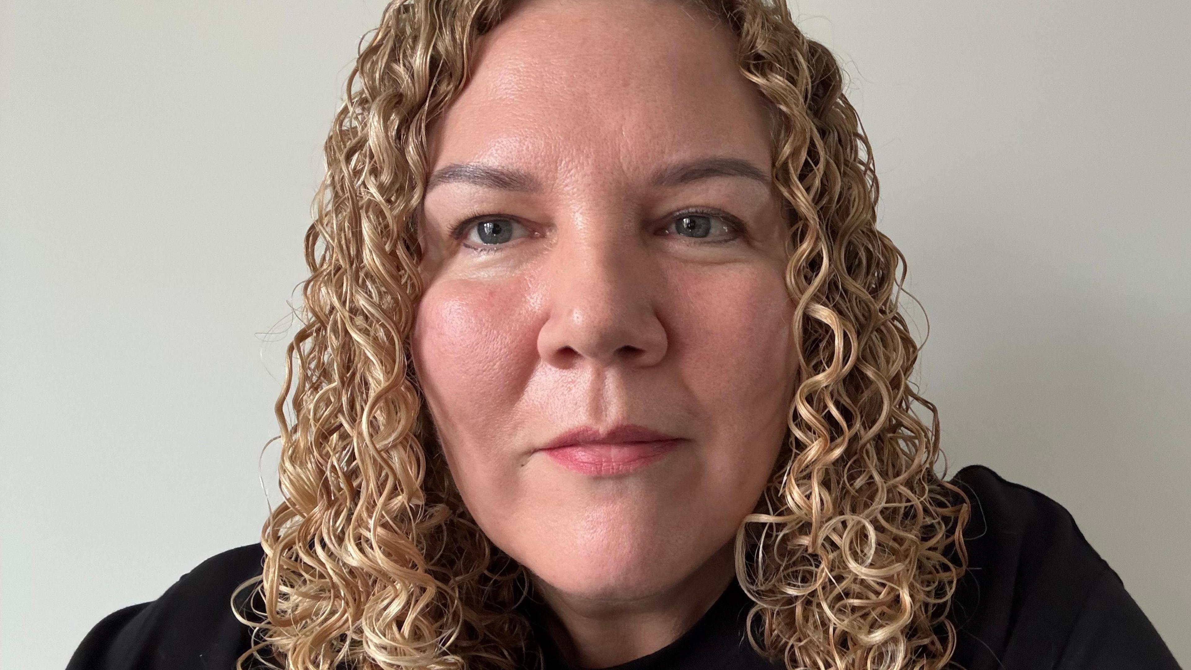 A woman with blonde curly hair looks straight to camera. She is wearing a black top.