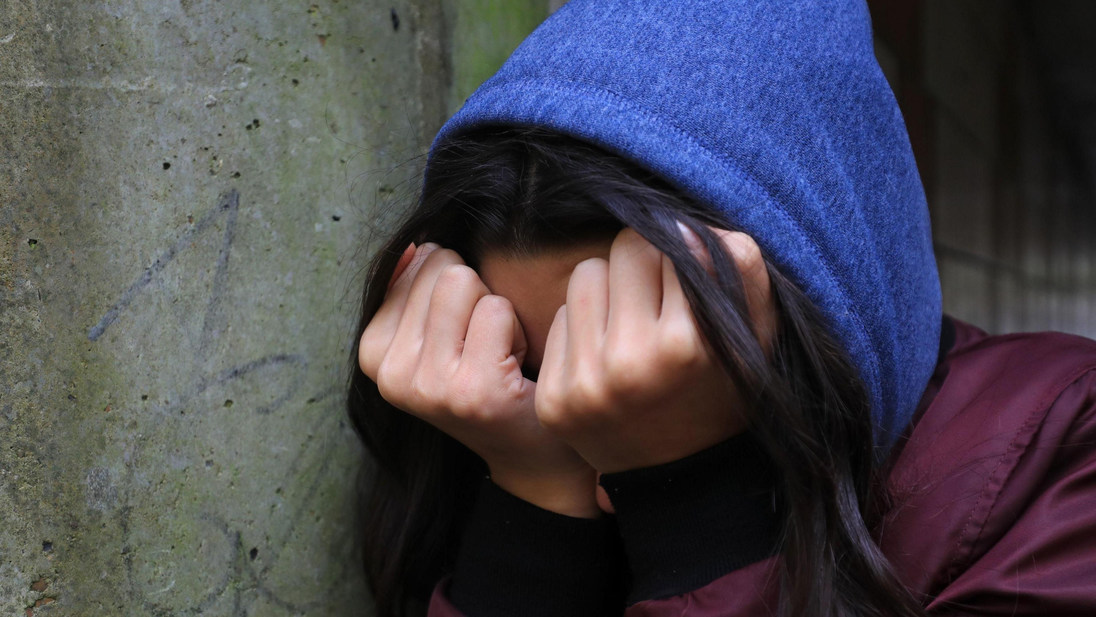 A teenage girl  wearing a blue hoodie and maroon coat  stands against a wall with her head in her hands .