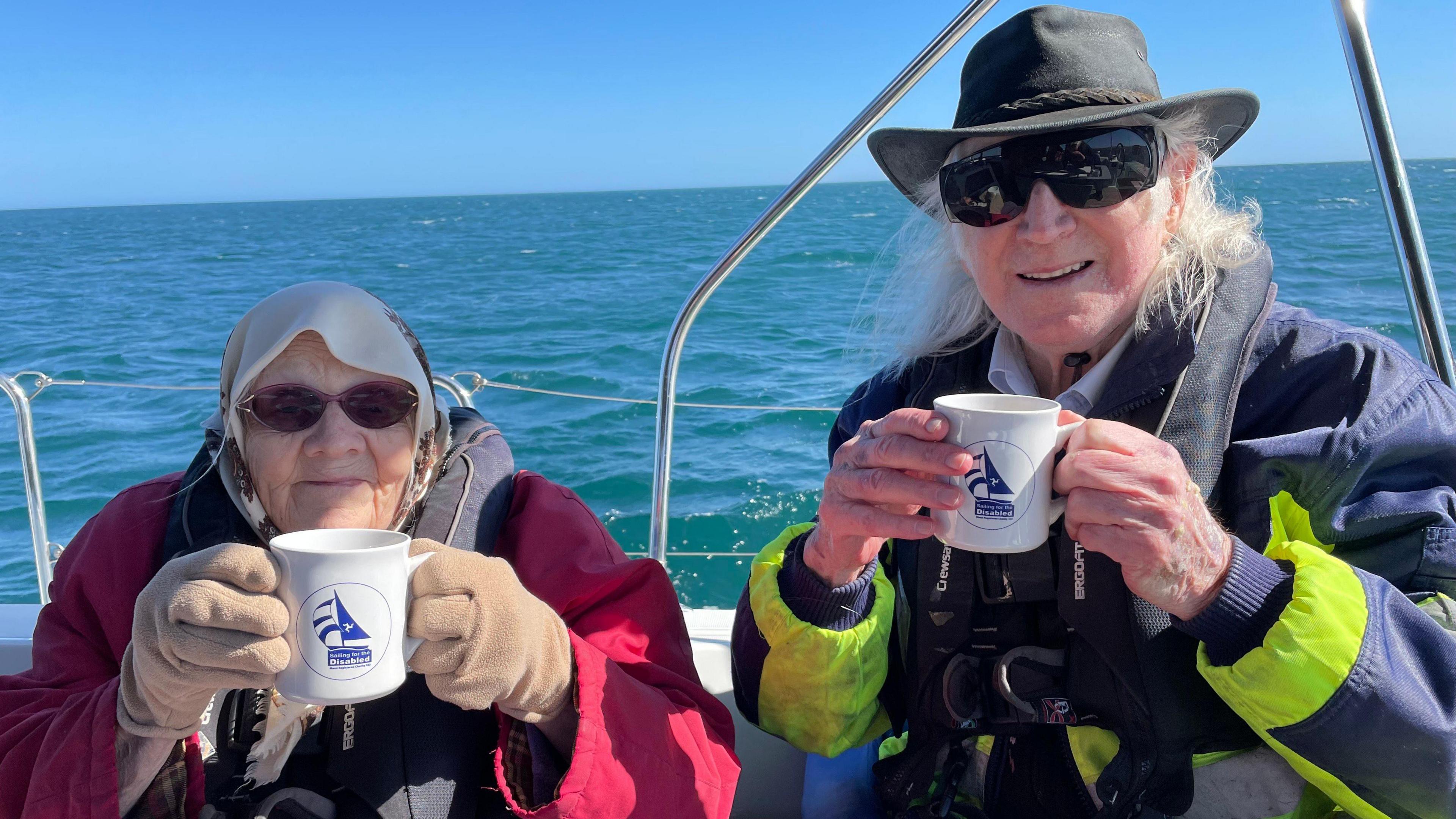 A woman wears a red coat, sunglasses and holds a white Sailing for the Disabled mug. A man sits next to her with long white hair and a hat. He also wears sunglasses and a bright jacket. The blue sea and cloudless skies are behind them on a calm day.