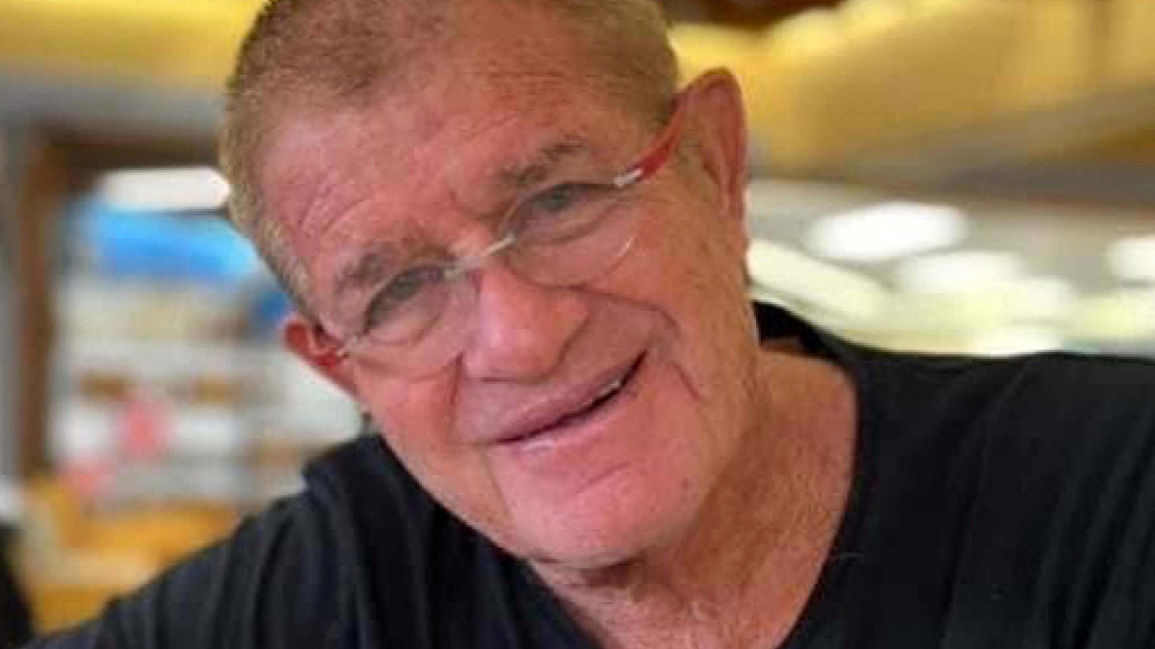 In this photograph taken when he was alive, Meny Godard, who has short light-coloured hair, glasses with red frames and is wearing a black t-shirt, smiles at the camera