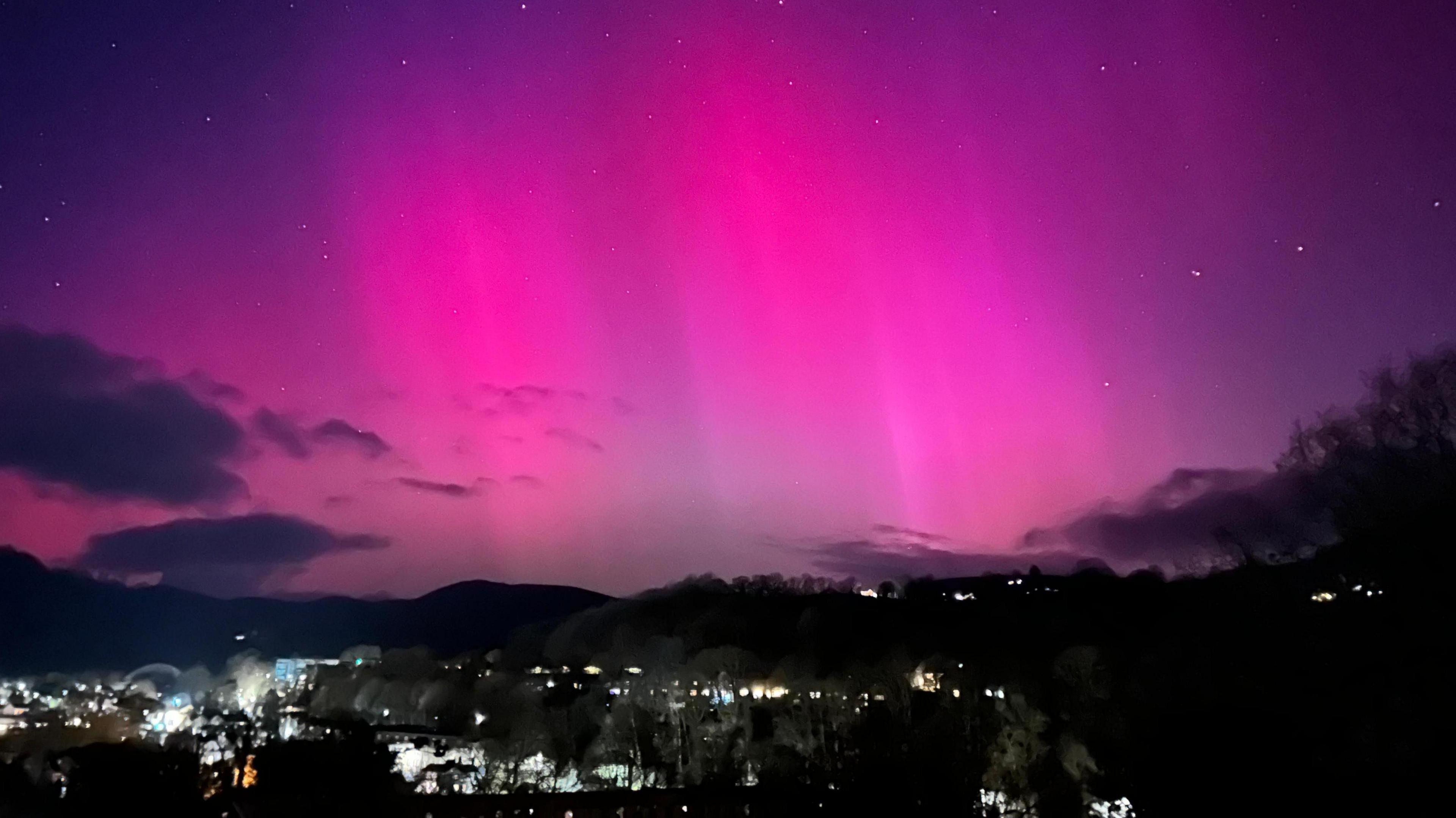 Vivid pink beams in purple starry skies above dark hills and illuminated town