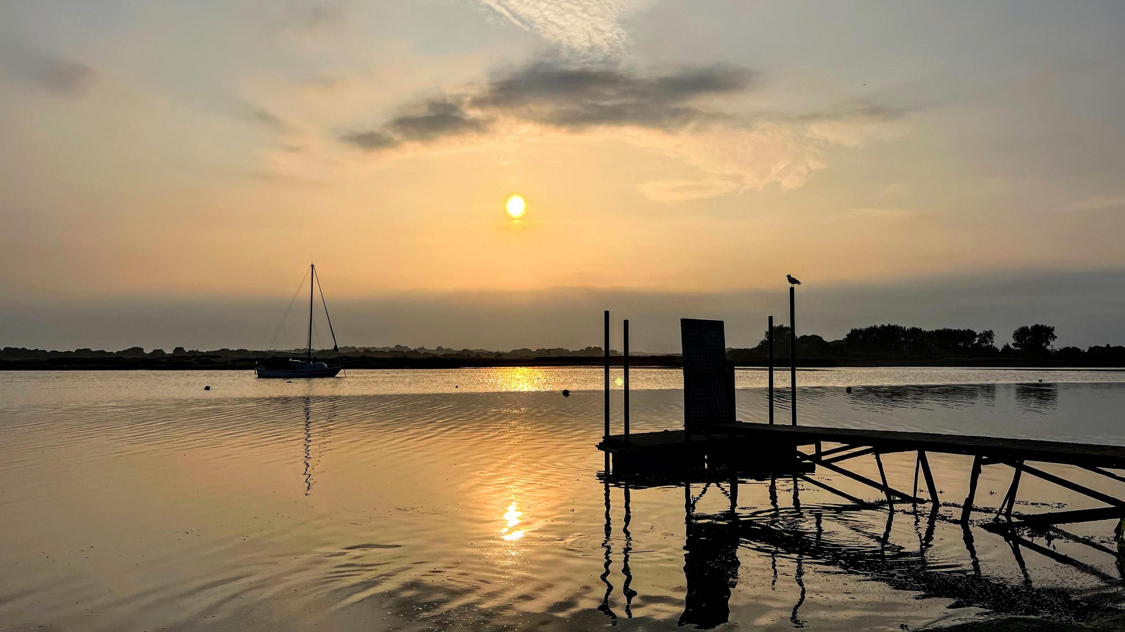 A low sun reflects on the water at Mudeford at sunset. A silhouette of a jetty can be seen in the foreground while a small sailing boat can be seen in the background. 