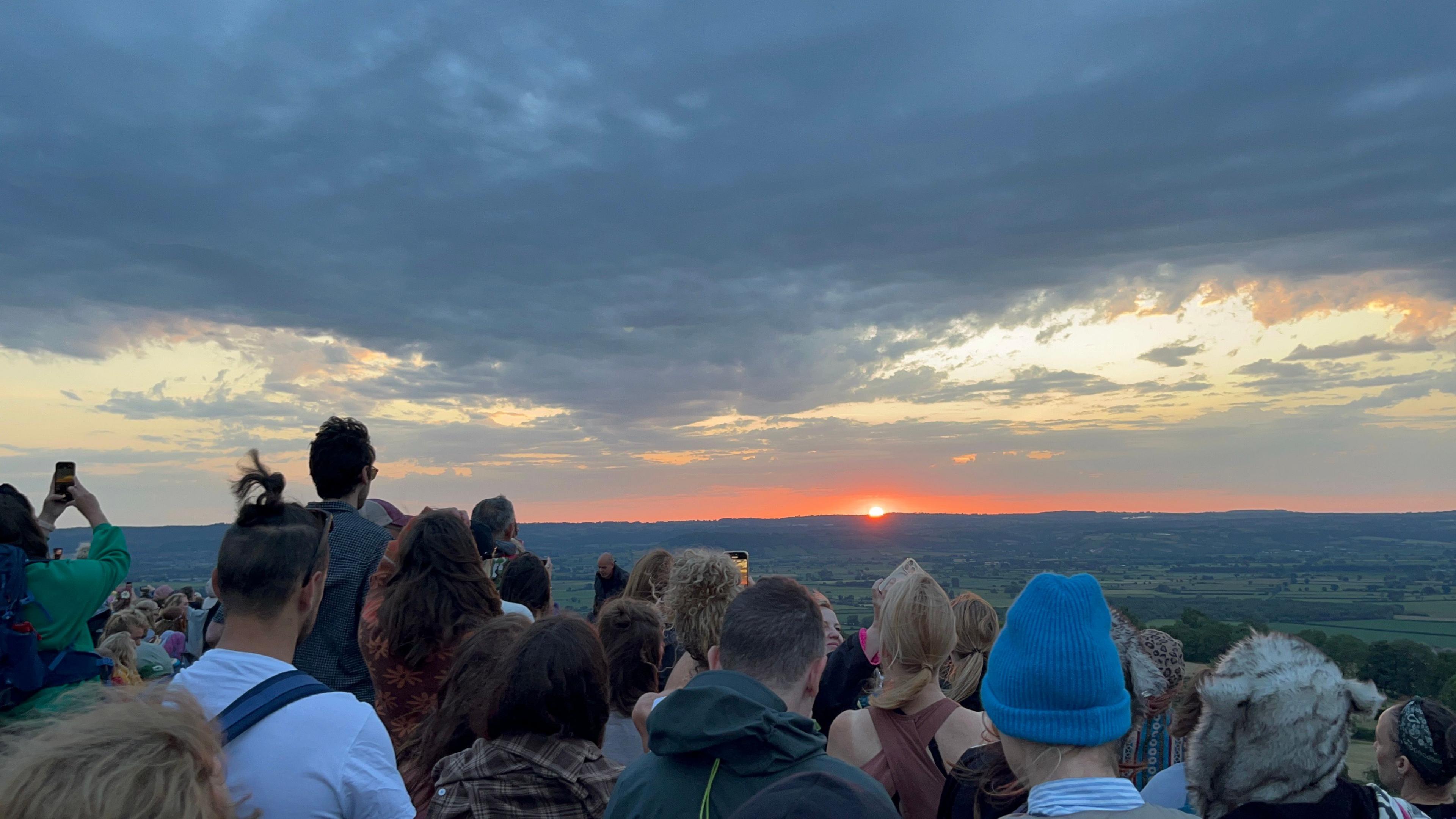 The sun rises in the distance as seen from Glastonbury Tor, the top of a hill. Around 20 people look out over fields in the distance. Some cloud is in the sky.