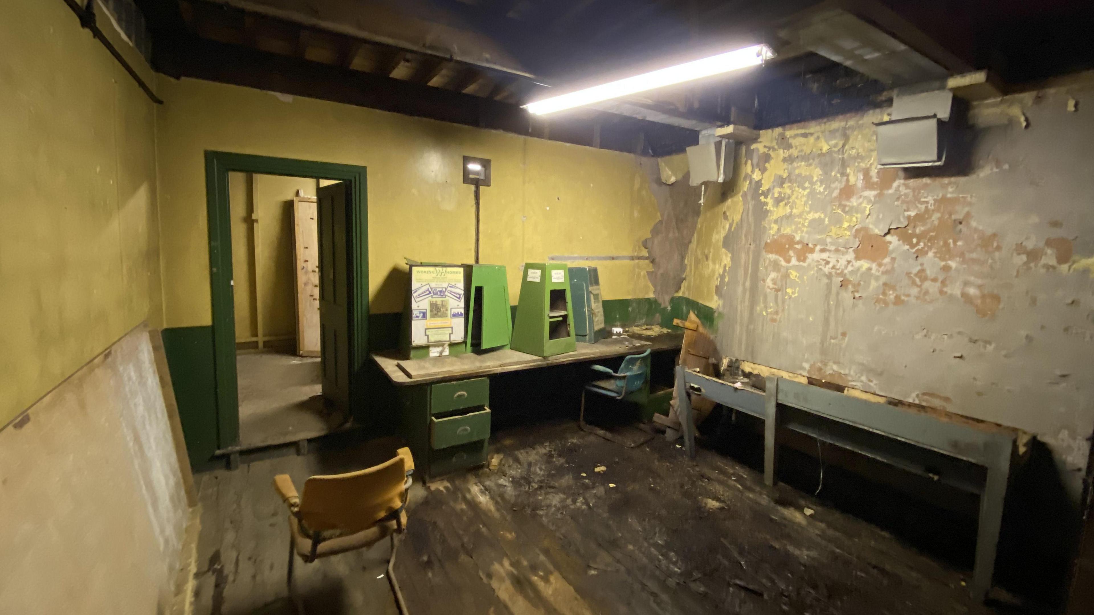 A small, windowless room. It is painted yellow, but paint is falling from the walls. There is a green desk and yellow chair in the room. The floor is dark wood. There is a single strip light.