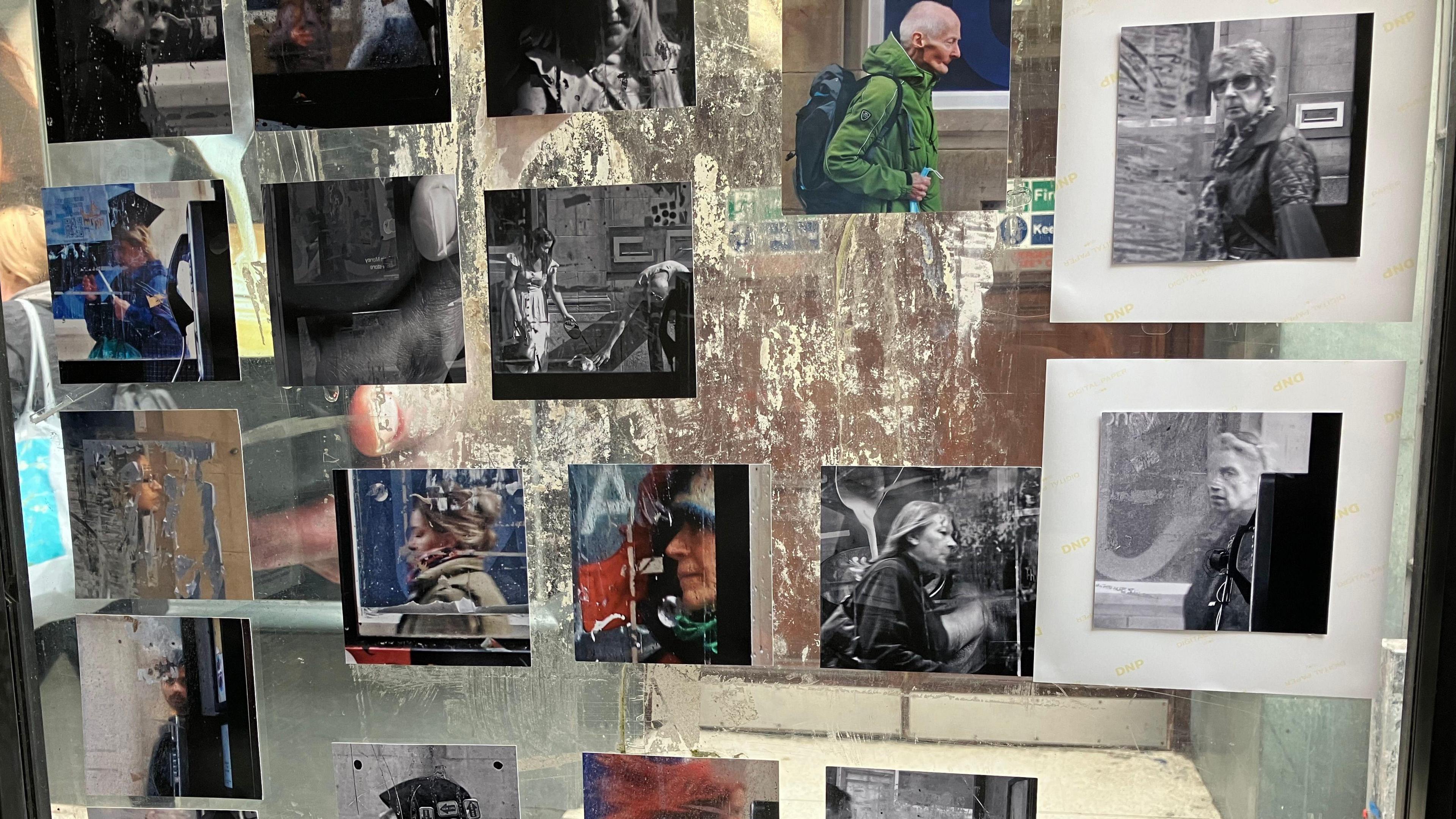 Several photographs are stuck the glass window of a phone box. They are a mixture of black and white and colour, and usually feature people walking by