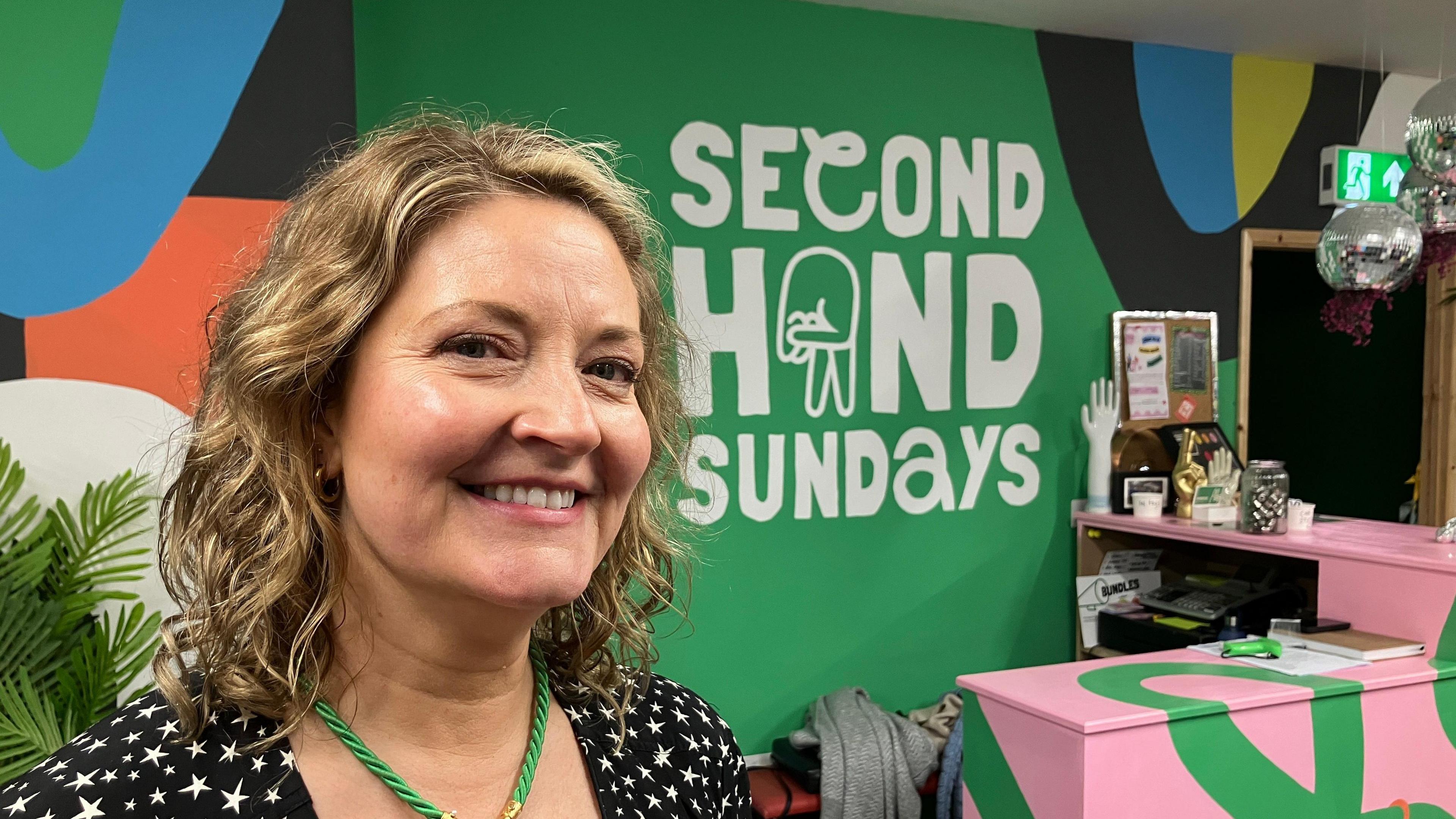 Alice Trethewey pictured smiling with shoulder-length dark blonde hair. She is wearing a black top with white stars and a green rope necklace. Behind her is a sign that reads Second Hand Sundays.