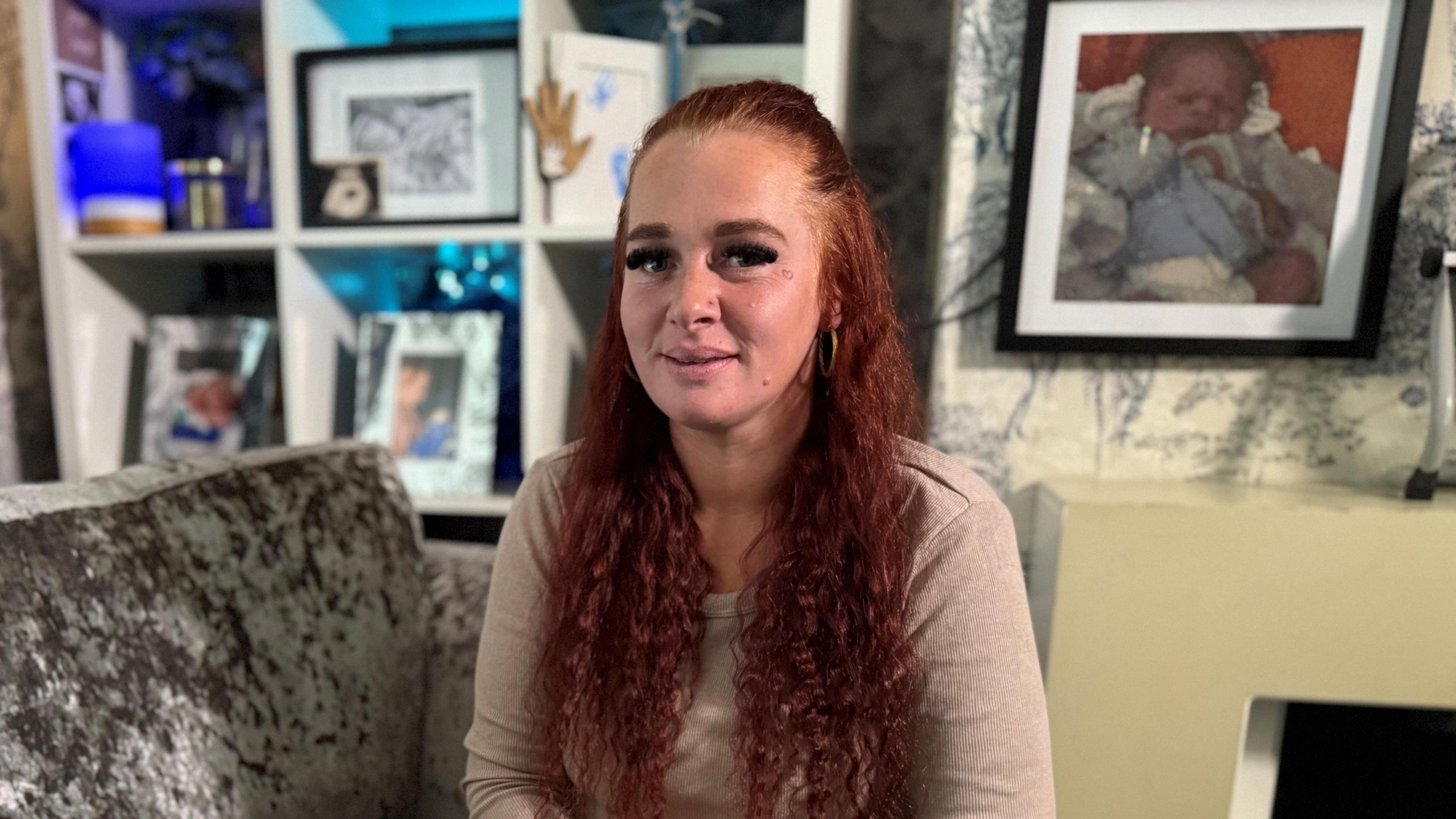 A woman with long wavy dark ginger hair tied back at the forehead wearing a pale pink long-sleeved top. She is sitting on a silver brushed velvet sofa. Behind her above her right shoulder is a white shelving unit with framed photos in it. Above her left shoulder is a large framed picture on the wall above a fireplace of a baby wearing a pale blue top.