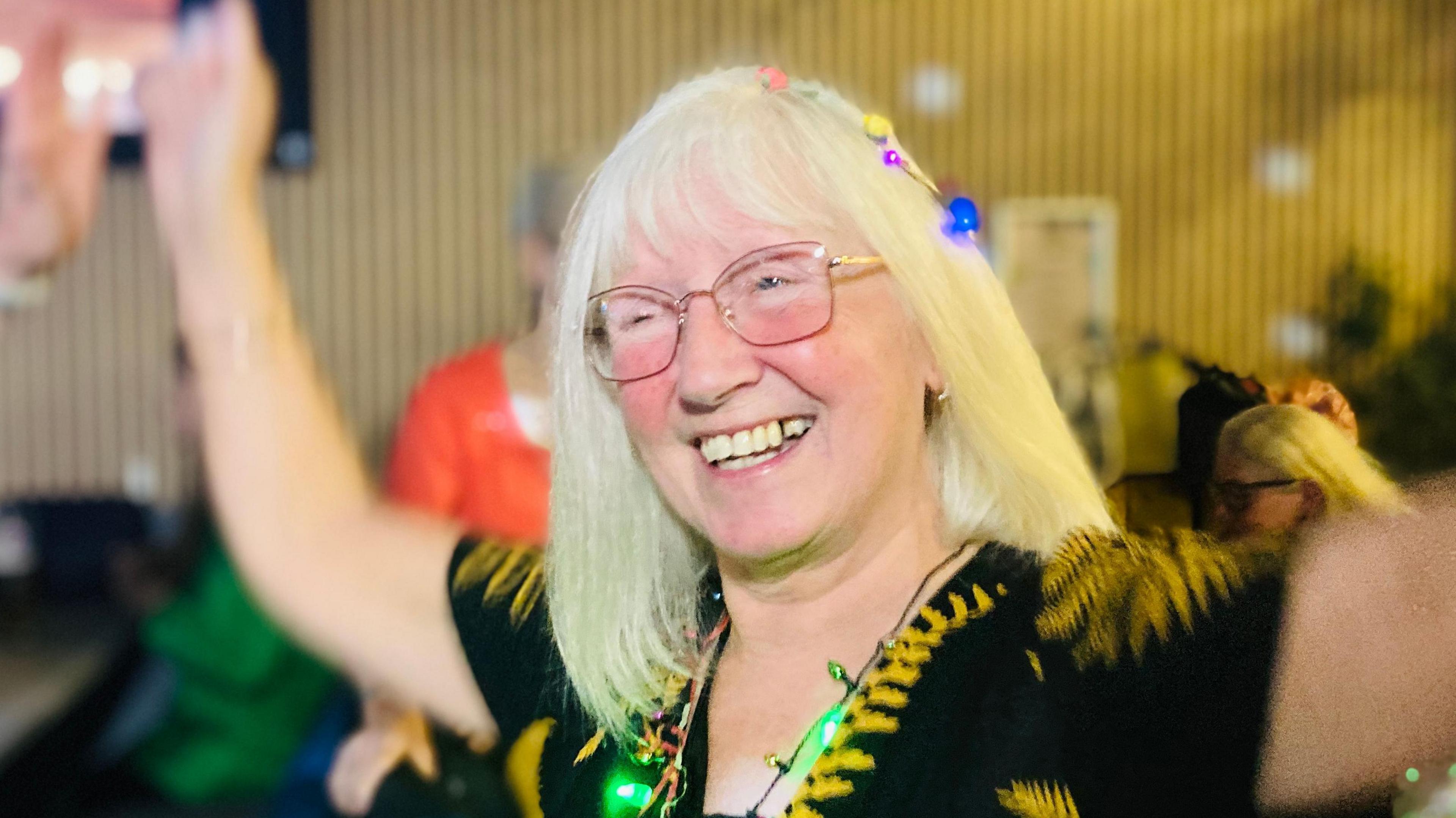 Lady dancing with her arms up in the air. She has fairy lights in her hair and around her neck - and she is laughing.    