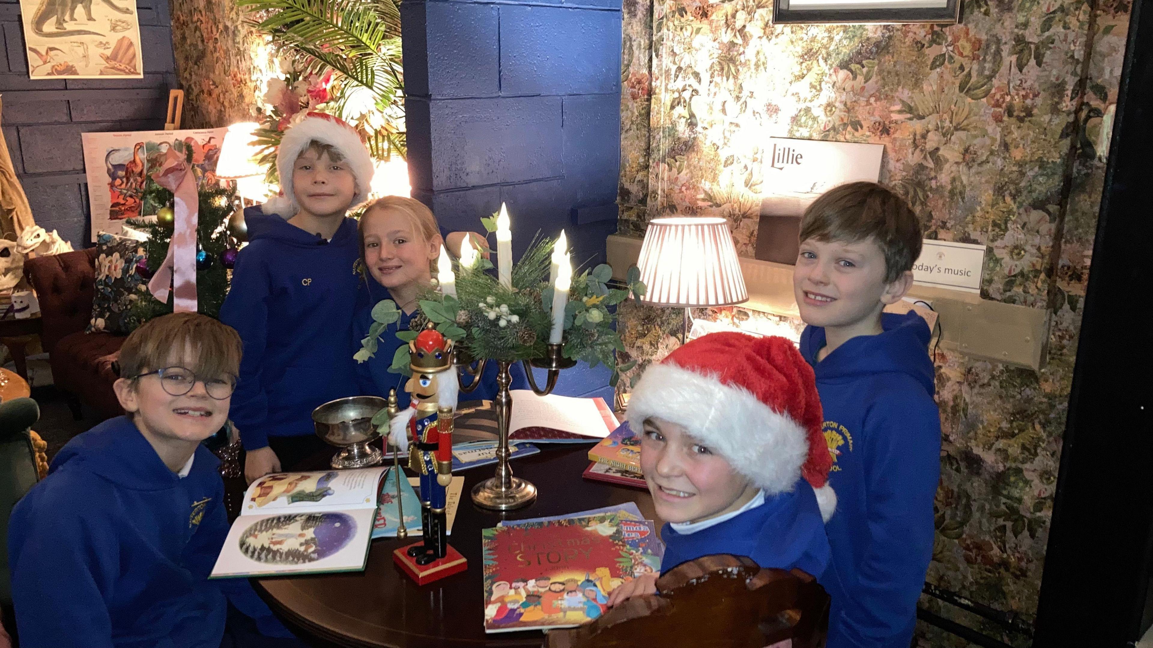 From left to right, Ollie, Christopher, Myles, Jesse and Joey sat around a wooden table with books and Christmas decorations on. There is floral decorations on the wall on the right and there is a blue wall on the left. The children are wearing blue school uniform and some are wearing Santa hats.