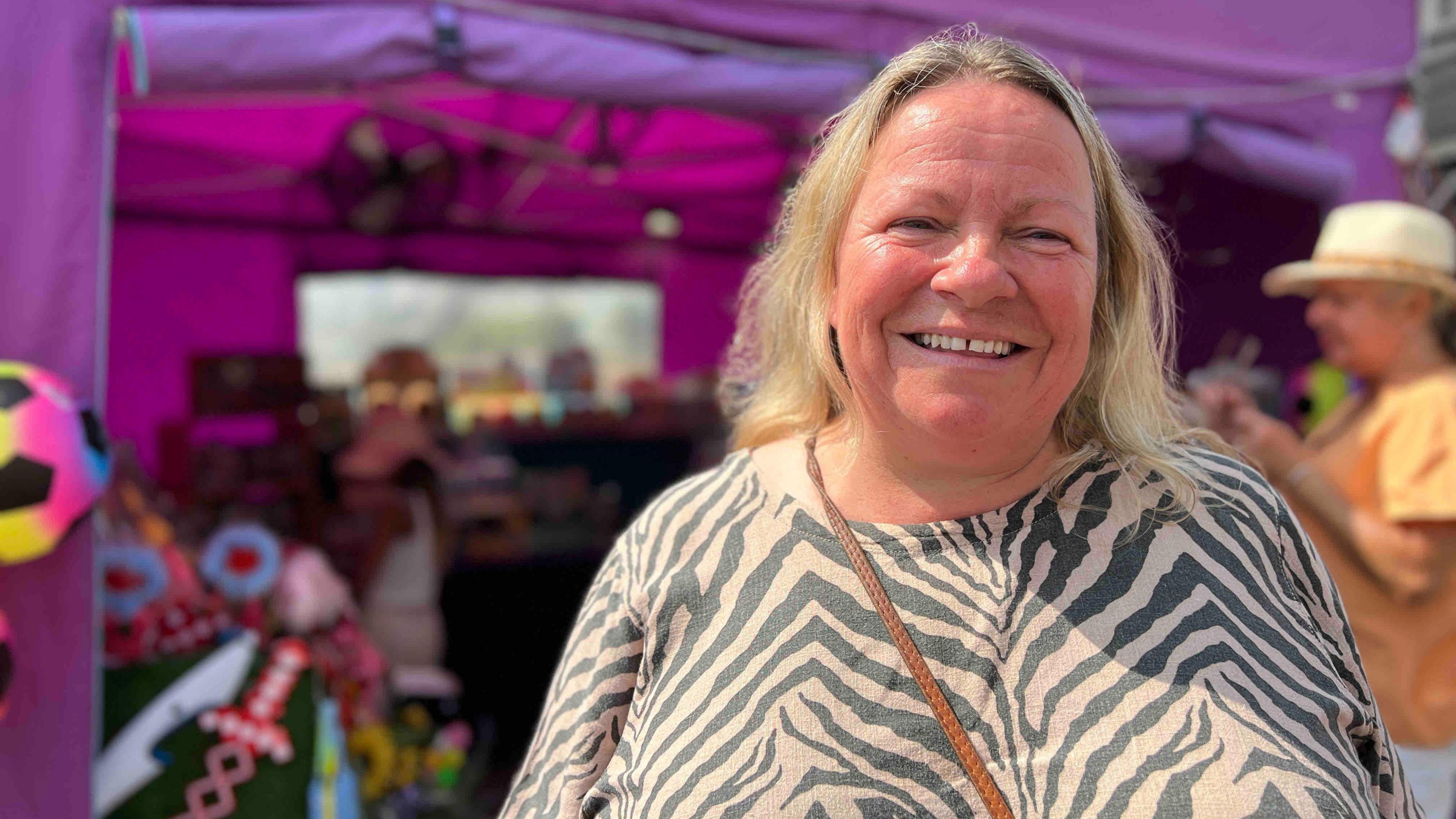 Kate is wearing a zebra-patterned top and she is standing in front of a purple tent. The tent contains colourful decorations and possibly toys or crafts. Other people are visible in the background under a clear sky. Kate has a brown strap across her chest, likely from a bag or purse.
