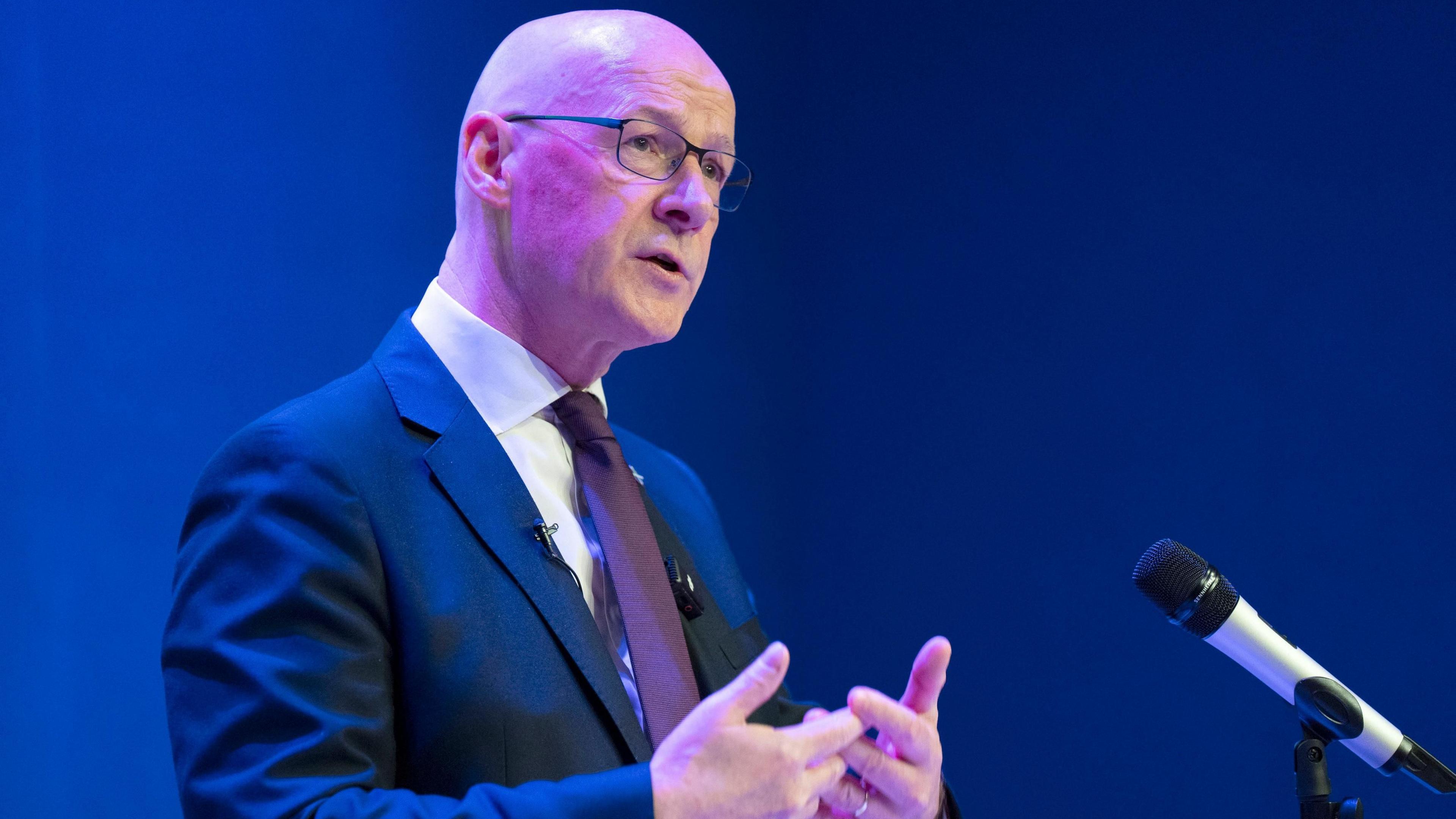 John Swinney standing on stage wearing a blue suit and a burgundy tie. There is a microphone to his right and there is a dark blue background.