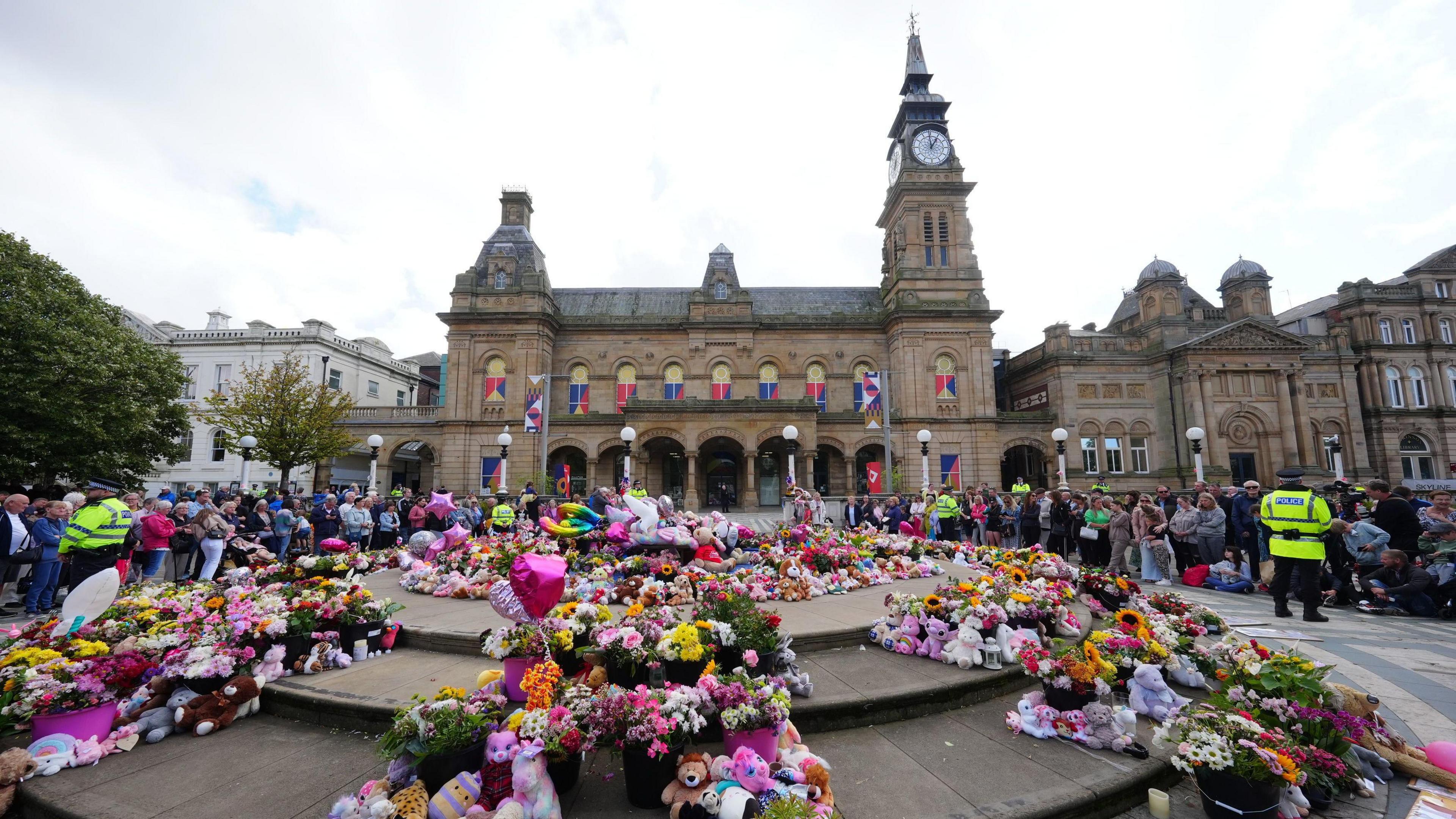 Rows of multi-coloured flowers outside the Atkinson Theatre, which is built with light-coloured stone and has a clock tower. People look on from the sides.