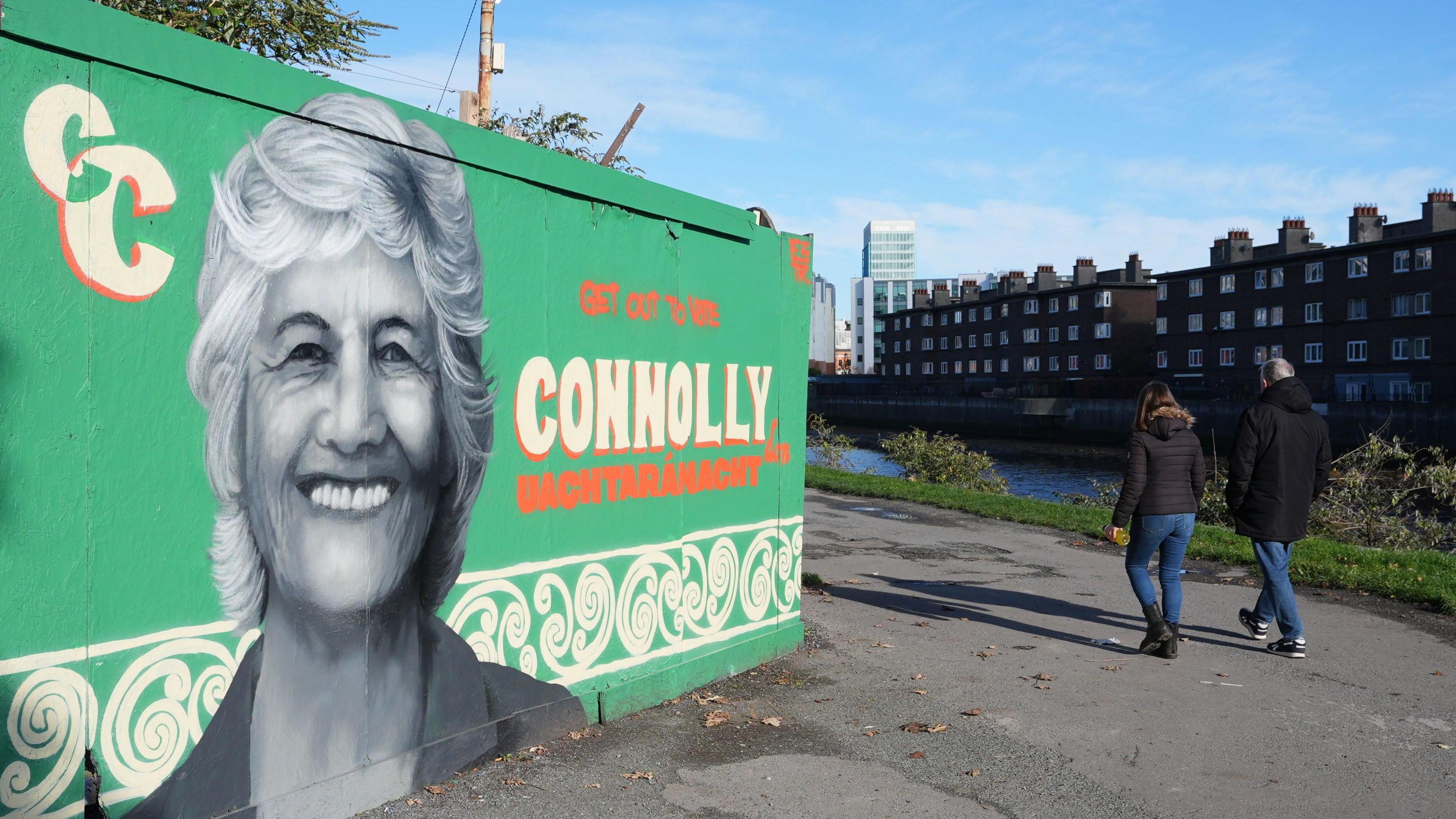 A black and white mural of a woman with short hair. It's on a green wall, which says Connolly in white writing with orange highlights. Two people are walking past the wall along a canal. Buildings can be seen in the background.