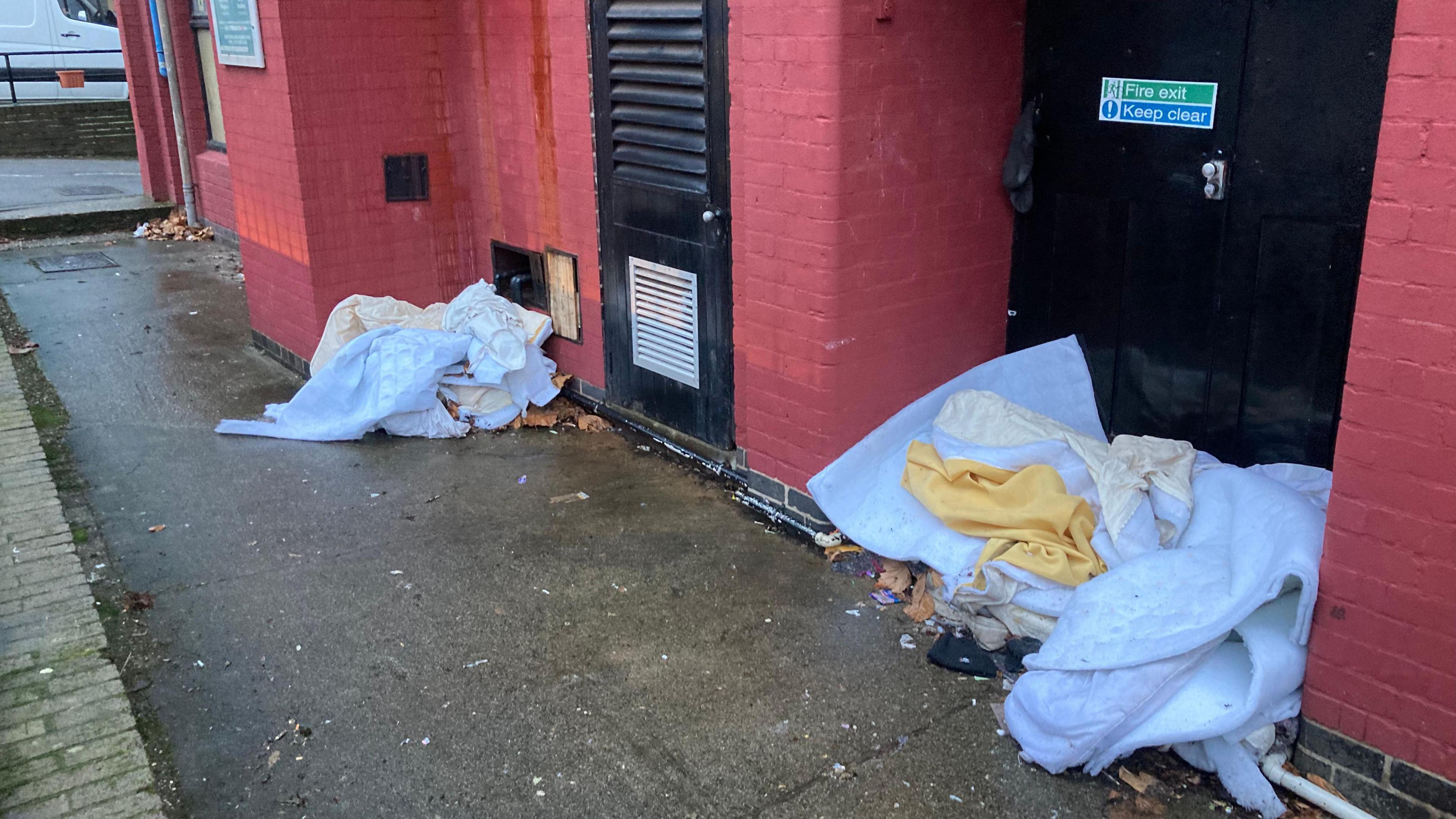 A pathway with white mattresses bundled up with various cloths on them showing rough sleeping on a street in Coventry outside a red brick-walled building.